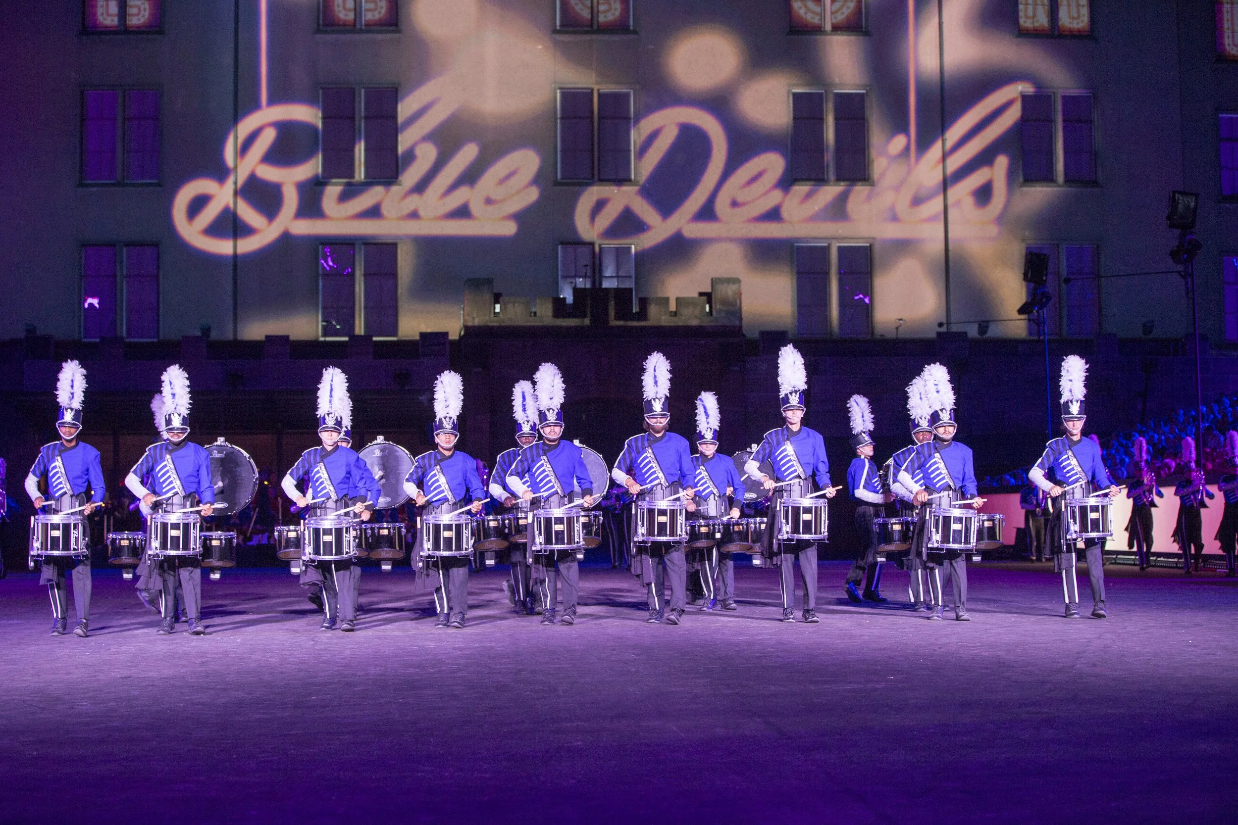 Eine Gruppe von Musikerinnen und Musikern in blauen Uniformen mit weißen Federschmuck auf den Hüten, die auf einem festlich beleuchteten Platz marschieren, im Hintergrund ist eine große Gebäudefassade mit dem projizierten Text 'Blaue Deltas' zu sehen.