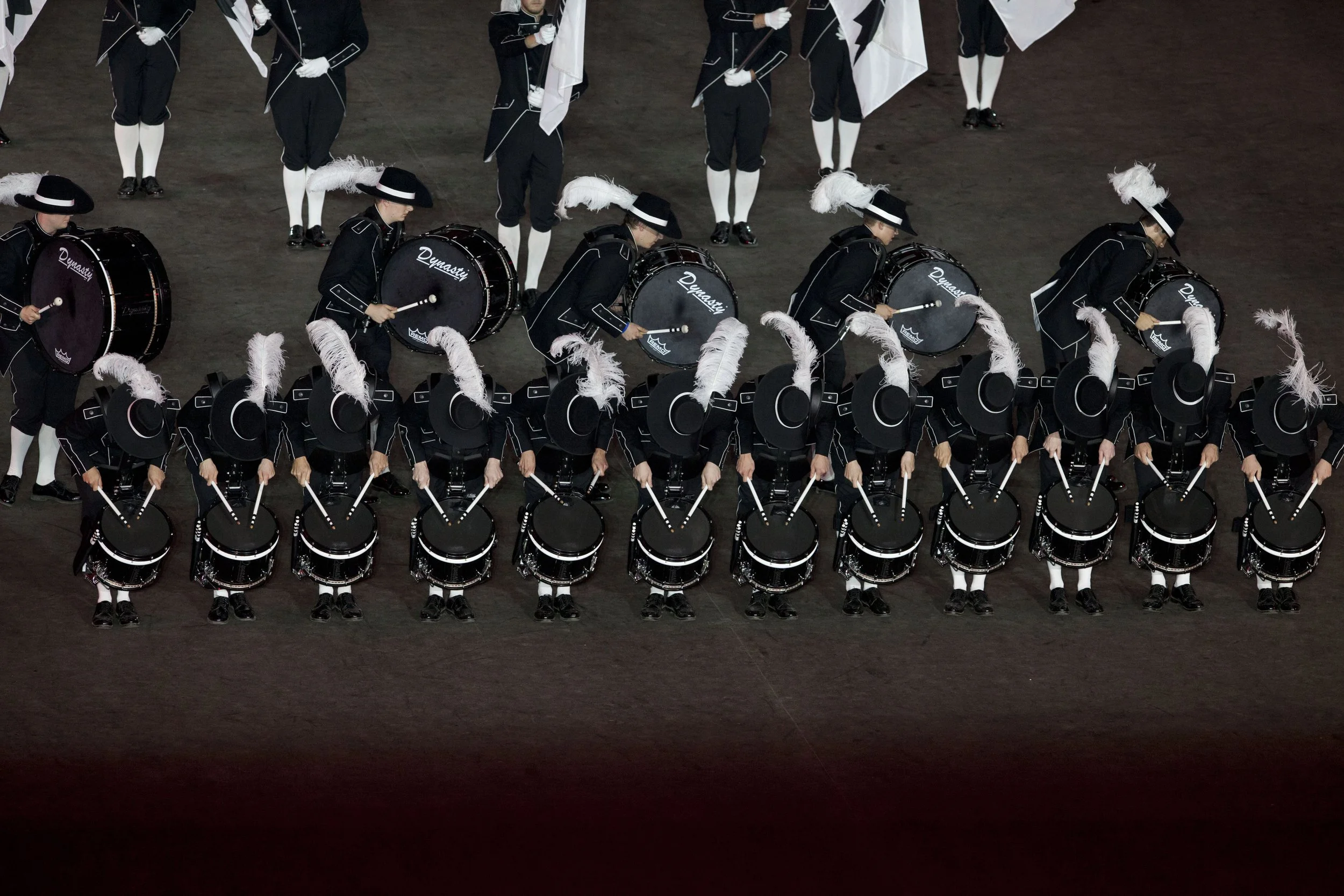 Eine Gruppe von marschierenden Musikern in schwarzen Uniformen mit weißen Federn auf Hüten, die auf dem Boden Trommeln spielen, während andere Marschkapellen im Hintergrund stehen.