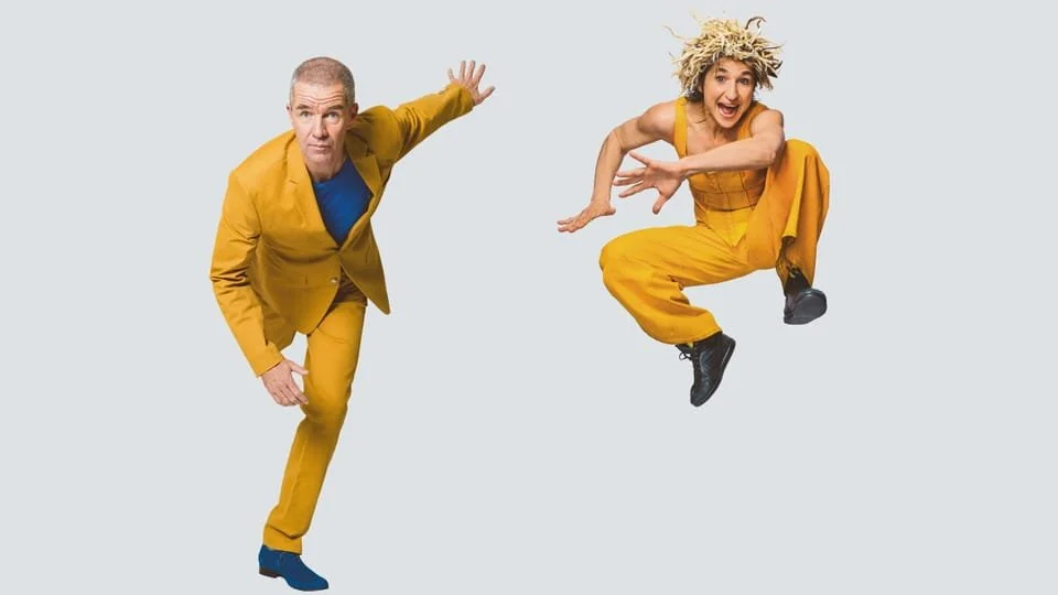 Zwei Menschen in gelben Outfits, die in der Luft springen und eine dynamische Pose einnehmen, vor hellem Hintergrund.