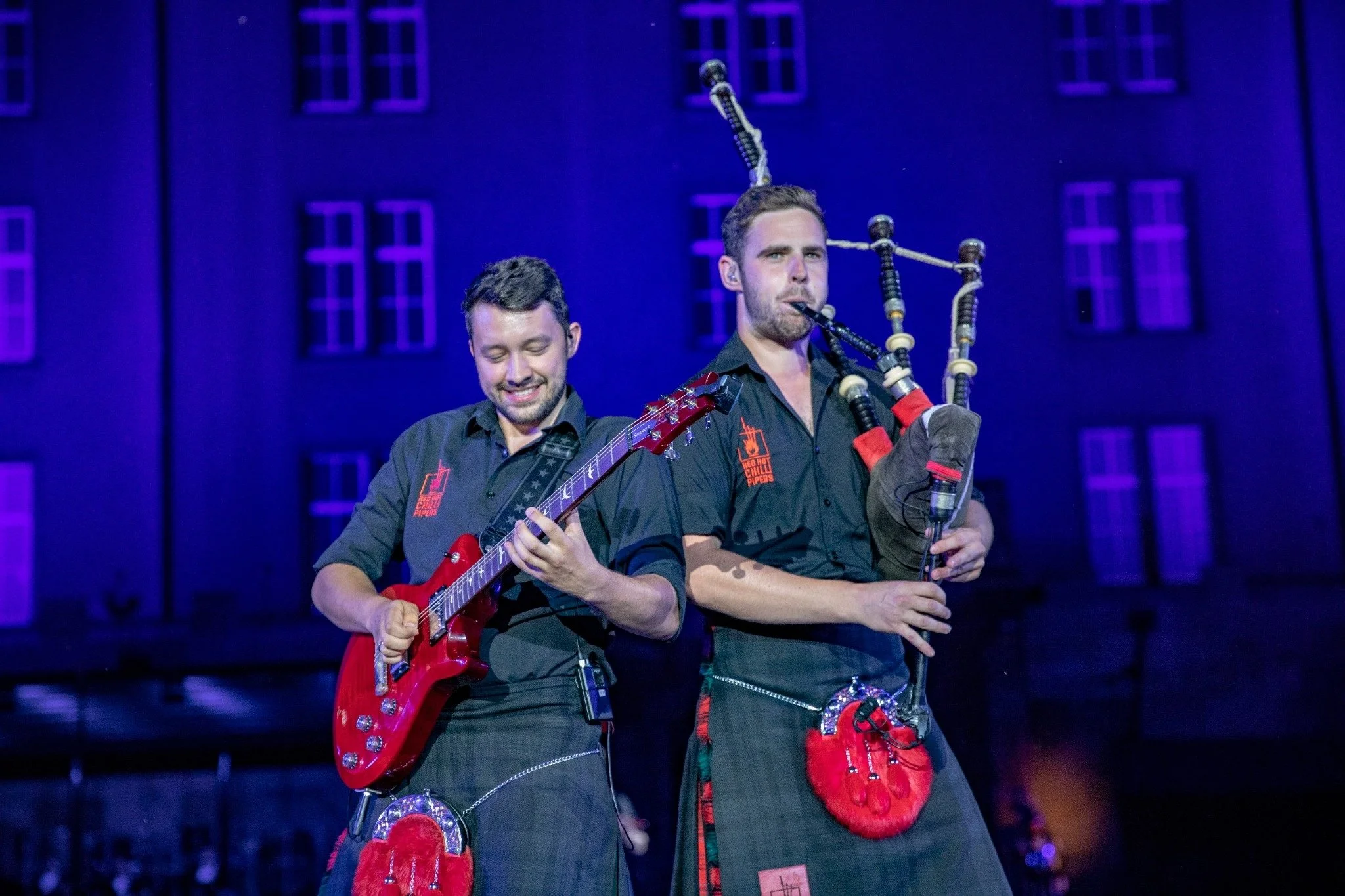 Zwei Männer, die an einem Theaterstück oder einer Show teilnehmen, tragen Kleidung im schottischen Stil und spielen Musikinstrumente, darunter eine Gitarre und Dudelsäcke, vor einer Bühne mit blauen Lichtern und Fenstern im Hintergrund.