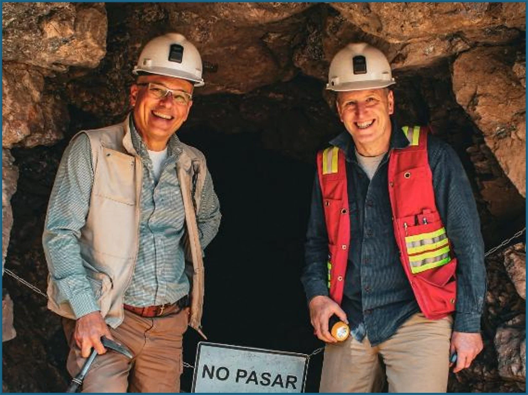 Two smiling men wearing helmets and outdoor gear standing in front of a cave. One is holding a flashlight, a 'No Pasar' sign is visible in front of them.