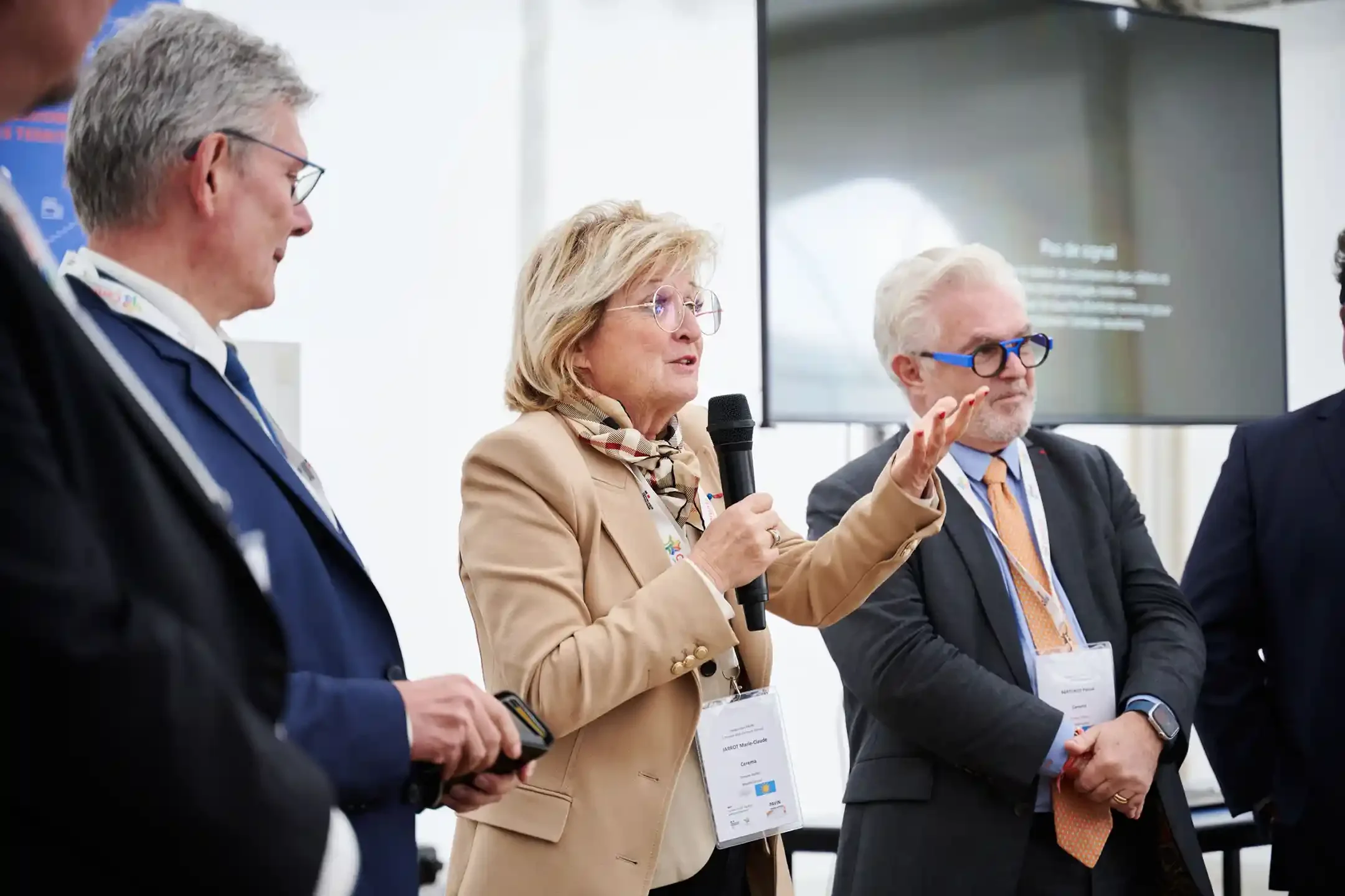 Une femme parle dans un microphone lors d'une conférence, entourée d'hommes en costume, devant un grand écran