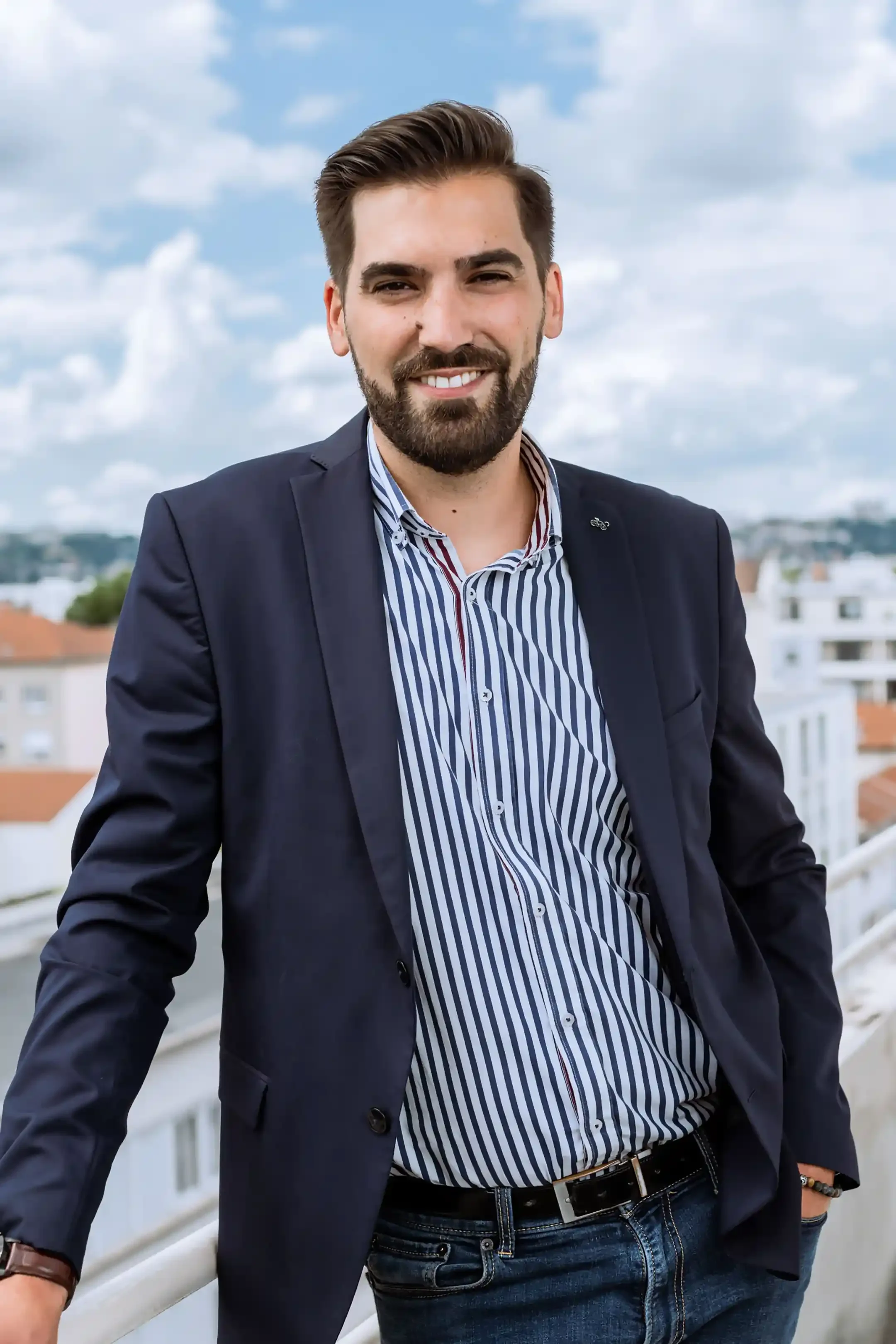 Un homme souriant, portant une veste noire et une chemise à rayures, debout sur un balcon avec un paysage urbain en arrière-plan, ciel nuageux.