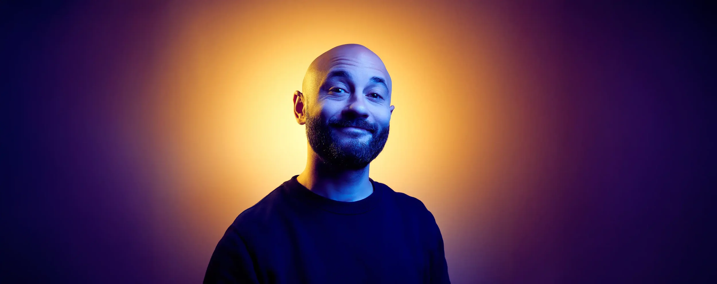 Homme souriant avec barbe et crâne rasé, fond coloré avec lumière jaune derrière sa tête