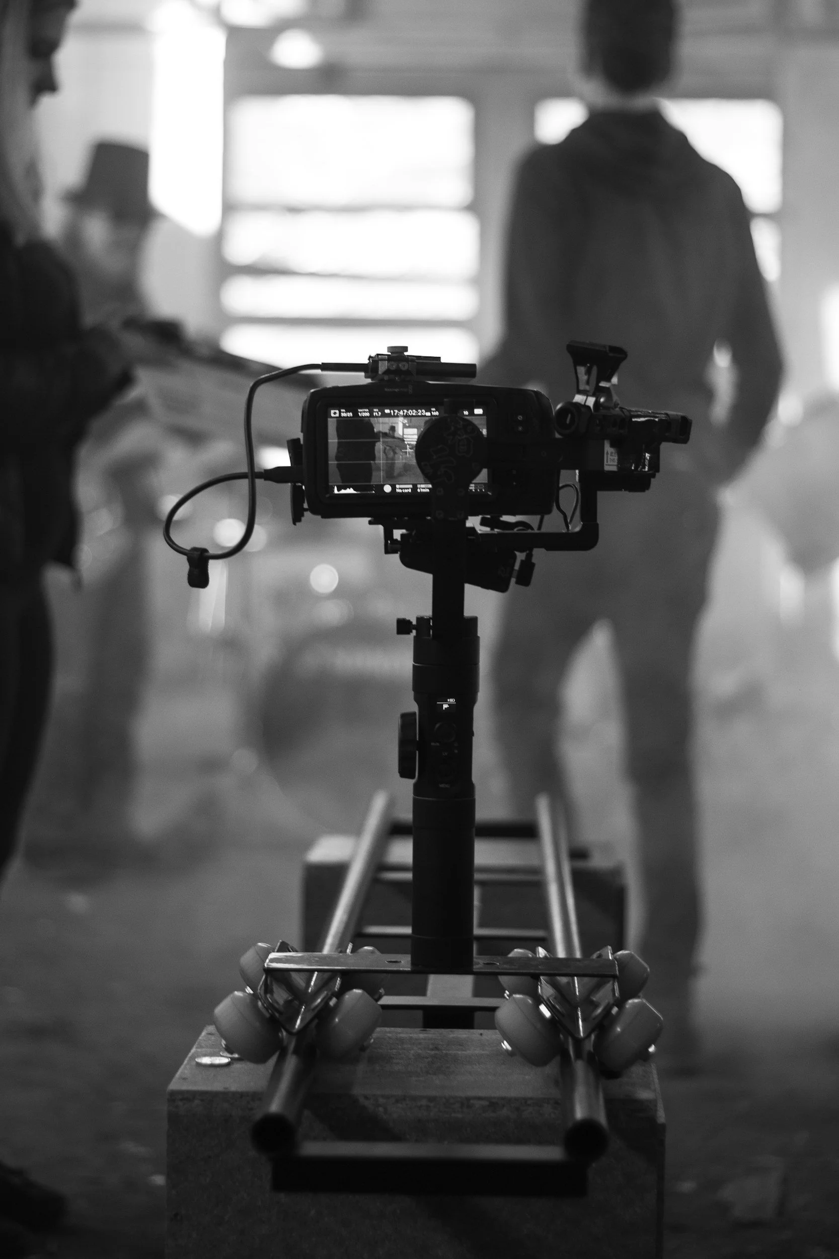 Une caméra de cinéma est placée sur un support à roulettes en studio, avec des personnes floues en arrière-plan, en noir et blanc.