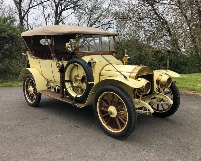 Voiture ancienne vintage de couleur beige avec détails en bronze, portant un toit en tissu beige, stationnée sur une route à côté d'un parc avec des arbres.