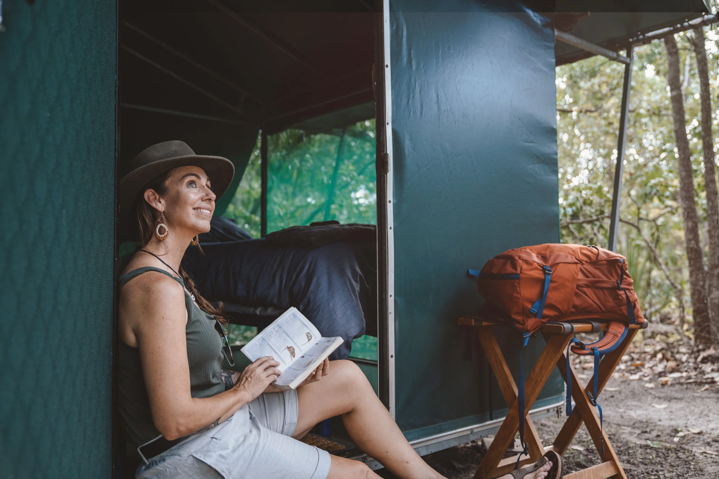 A woman sitting outside a camper van, reading a book, with a backpack and camping chair nearby, surrounded by trees.