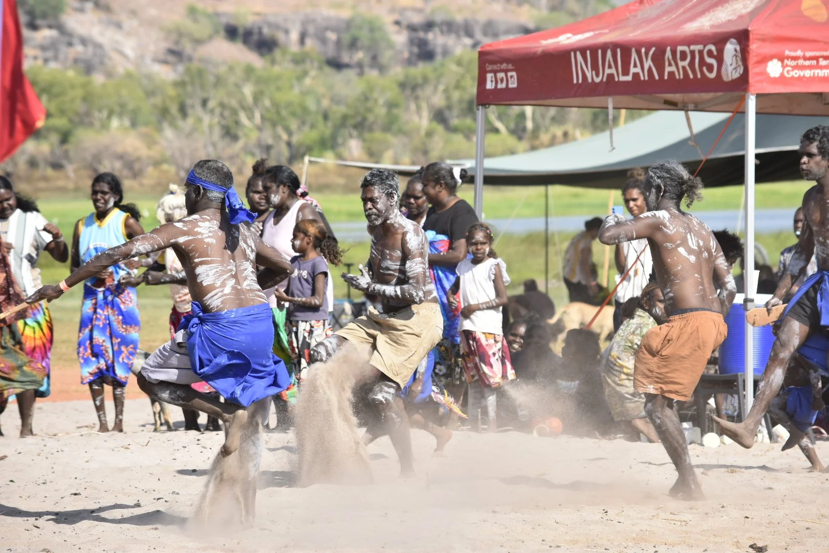 People participating in a traditional dance or celebration outdoors, with some covered in white paint, under a tent labeled 'INJALAK ARTS', with onlookers in the background and a rural landscape.