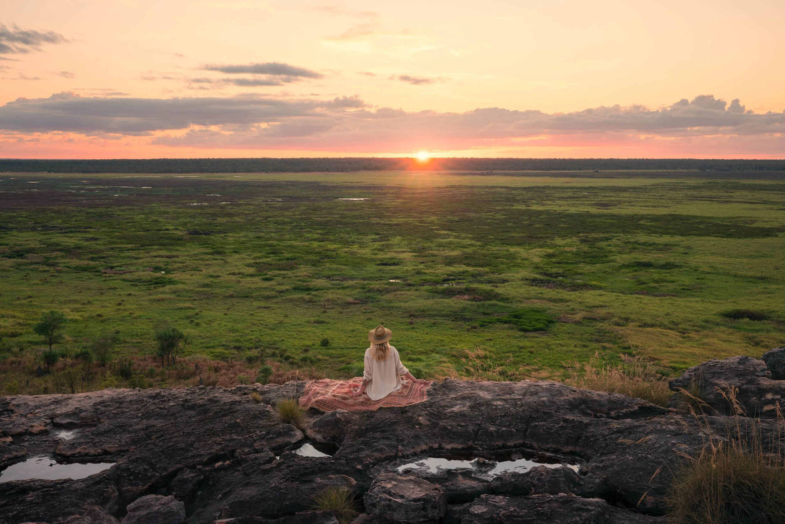 A woman sitting on rocks, facing away, watching the sunset over a vast green wetland landscape.