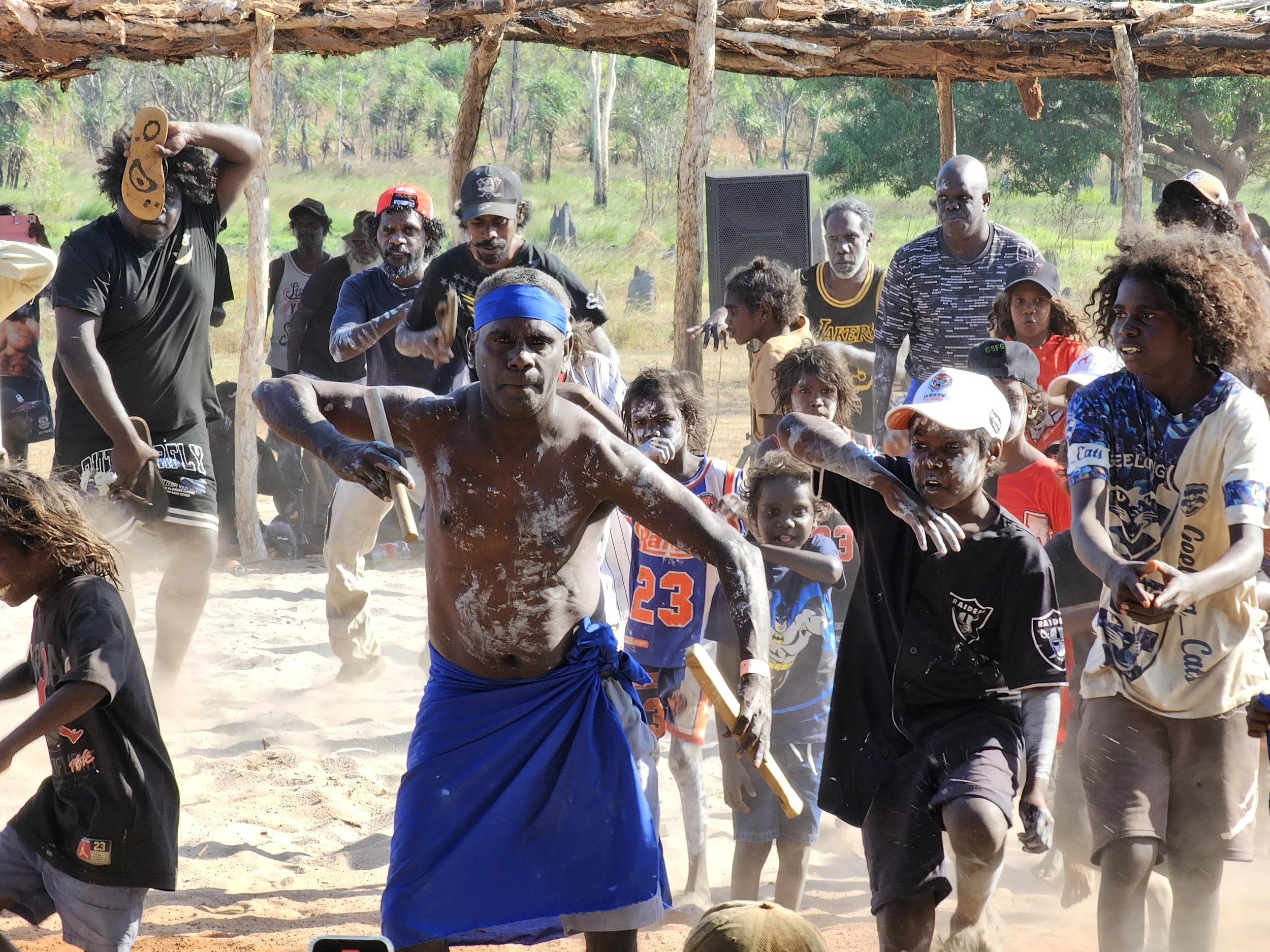 A group of people, including men, women, and children, participating in a traditional stick fighting event outdoors under a makeshift wooden shelter surrounded by trees and grassy land.
