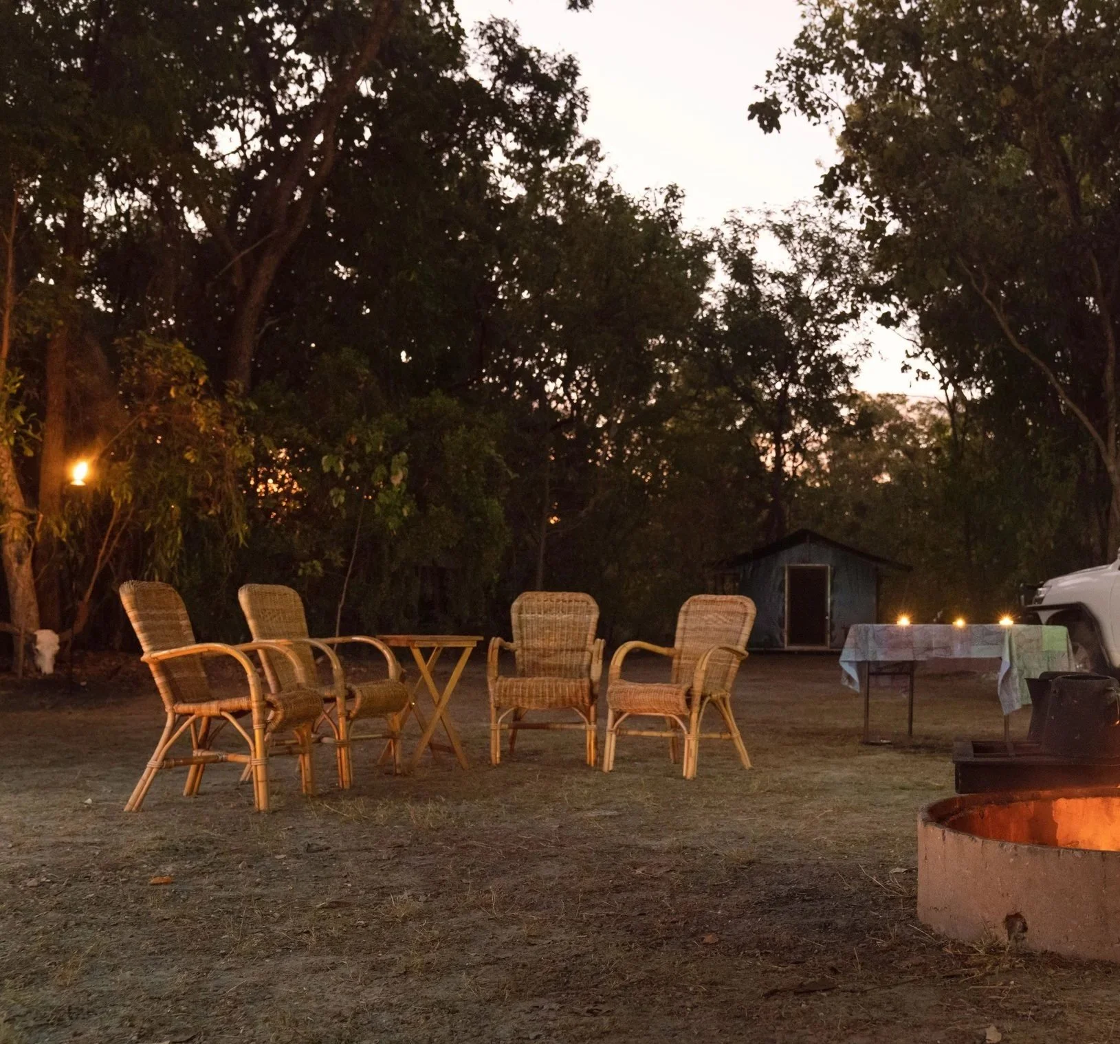 Outdoor patio area with five wicker chairs around a small table, a larger table with a tablecloth, a small shed in the background, and trees at dusk with a fire pit in the foreground.
