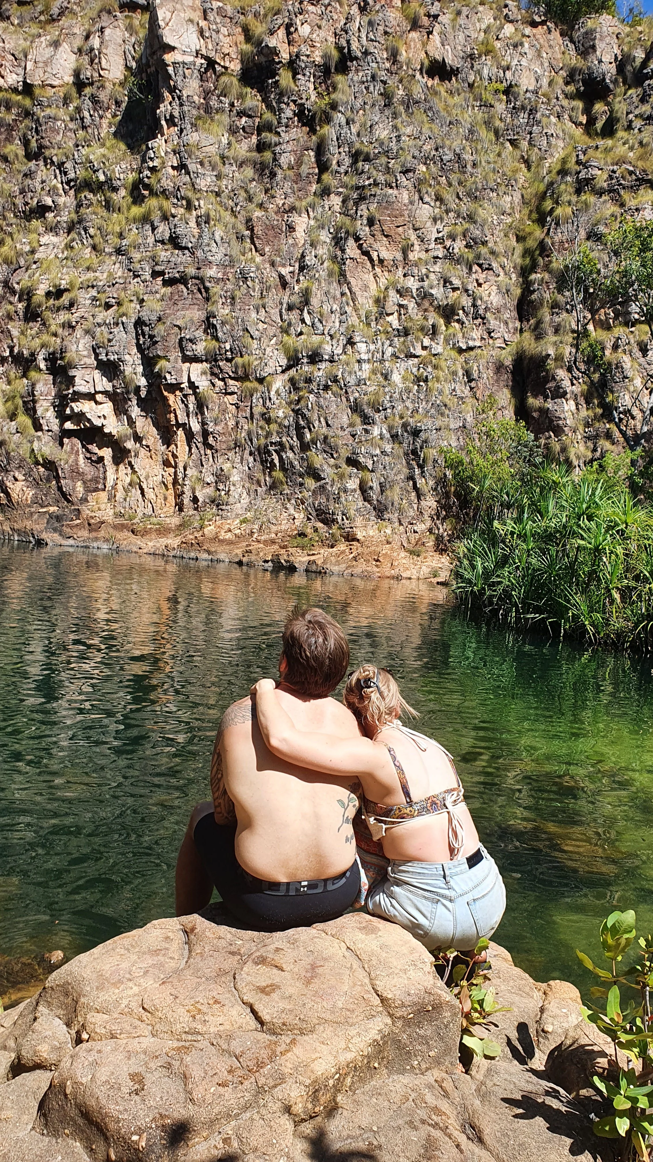A couple sitting on rocks by a body of water, with a rocky cliff and green plants in the background.