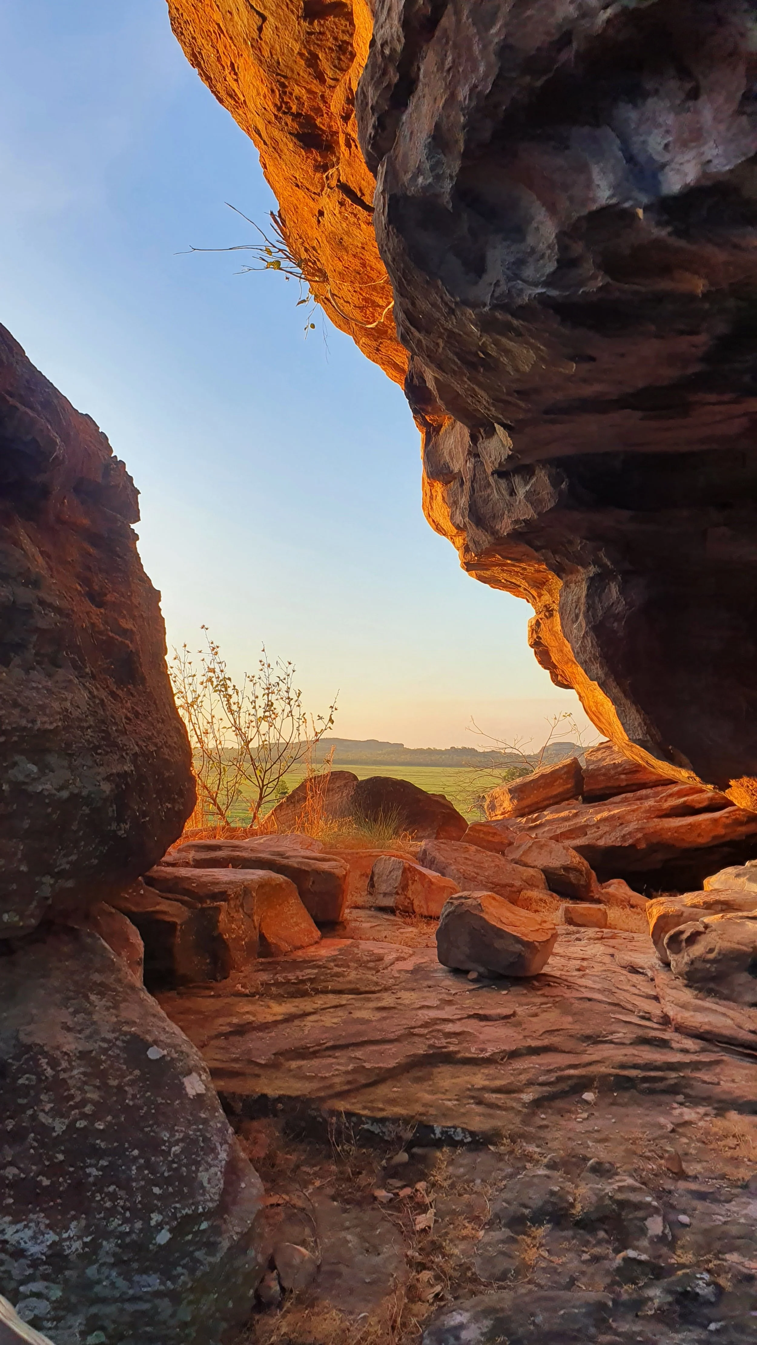 View through a rocky opening at sunset, with warm orange light illuminating the rocks and a landscape with sparse trees and distant hills visible in the background.