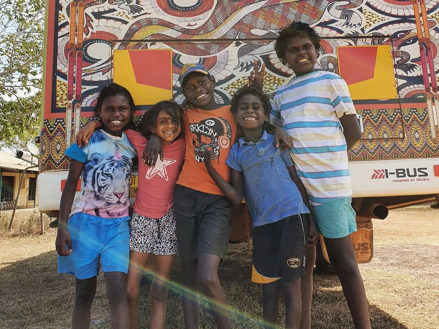 Six children standing together in front of a colorful decorated bus, smiling and showing gestures of happiness.
