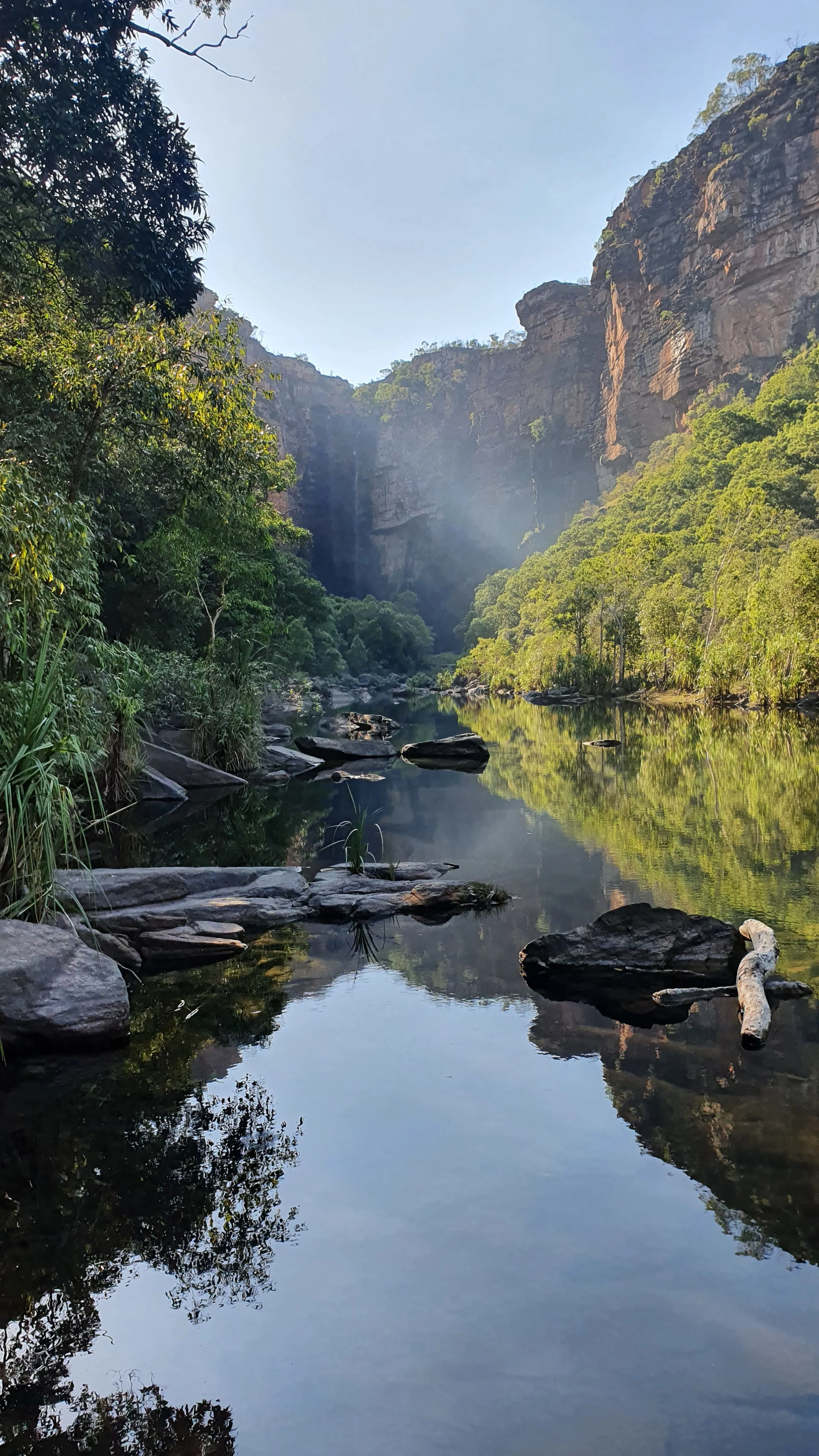 A peaceful river flowing through a lush green valley with tall cliffs and a waterfall in the background, under a clear blue sky.