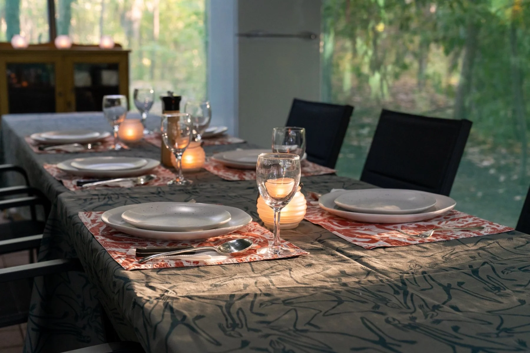 A dining table set for six with plates, silverware, wine glasses, and candles, in a room with large windows showing a green forest outside.