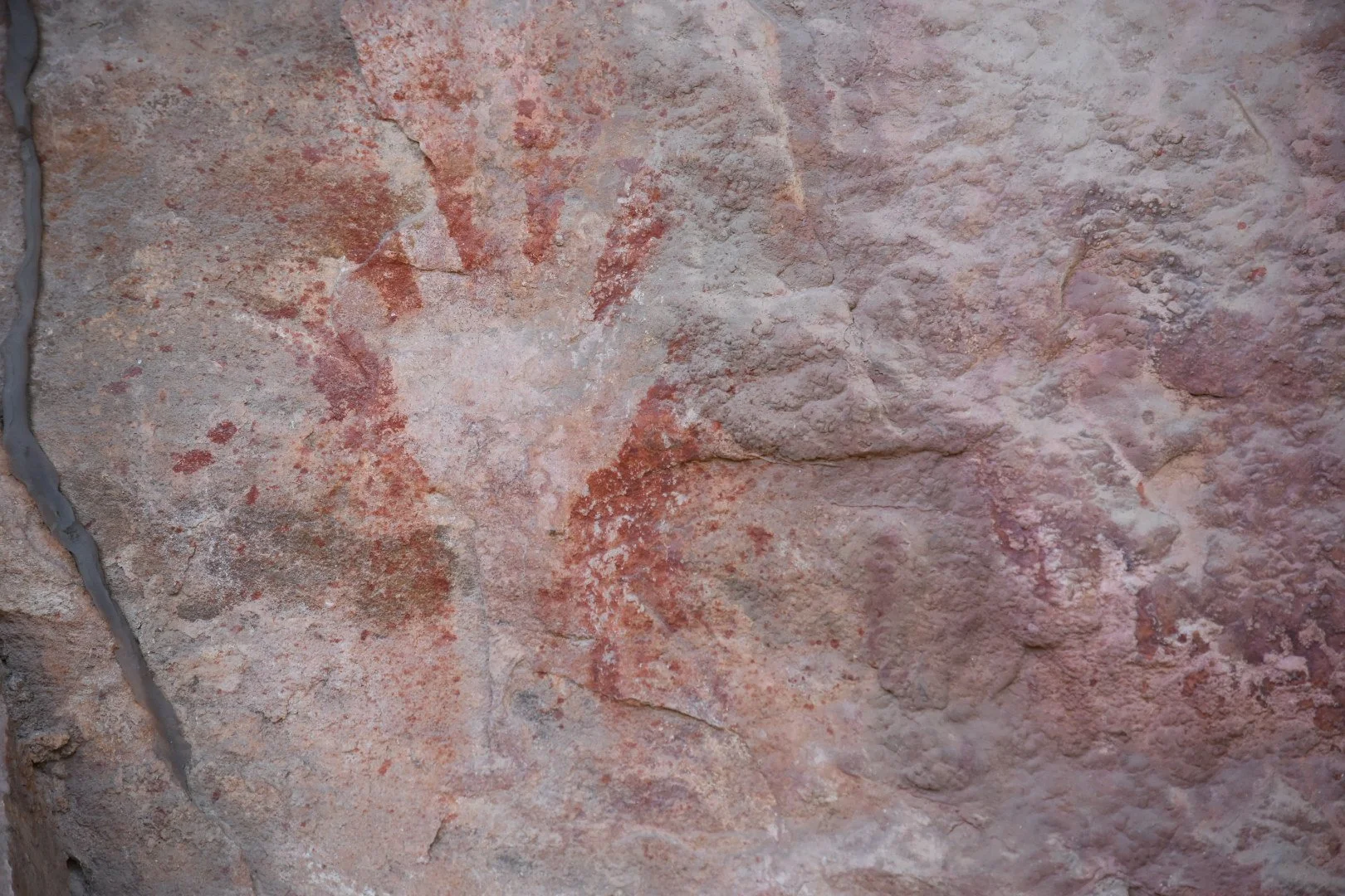 Close-up of a weathered, textured stone wall with reddish and beige hues, visible cracks, and some mortar along the left edge.