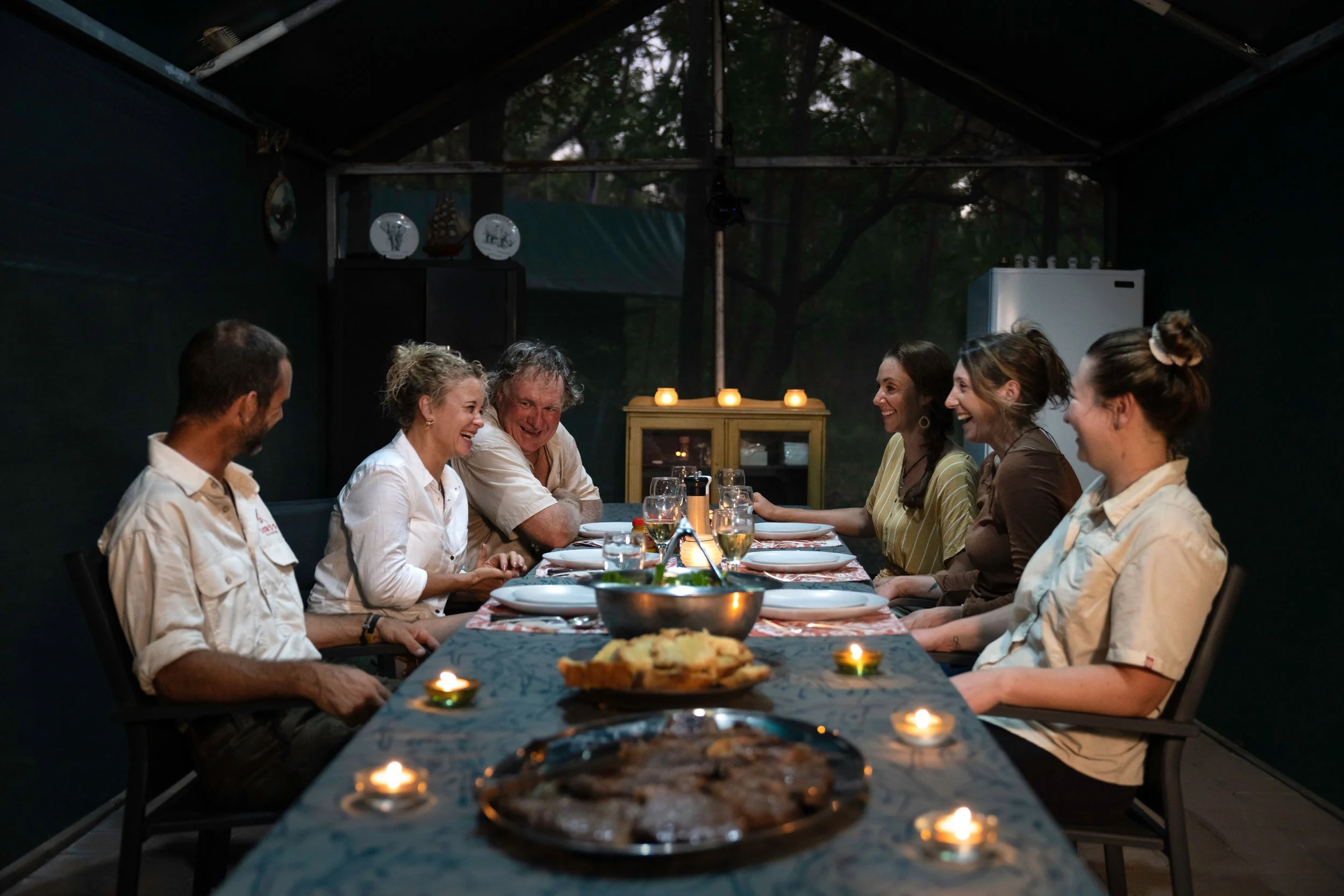 A group of six people sitting around a dinner table with food and drinks, smiling and enjoying conversation in a dimly lit outdoor setting at night.