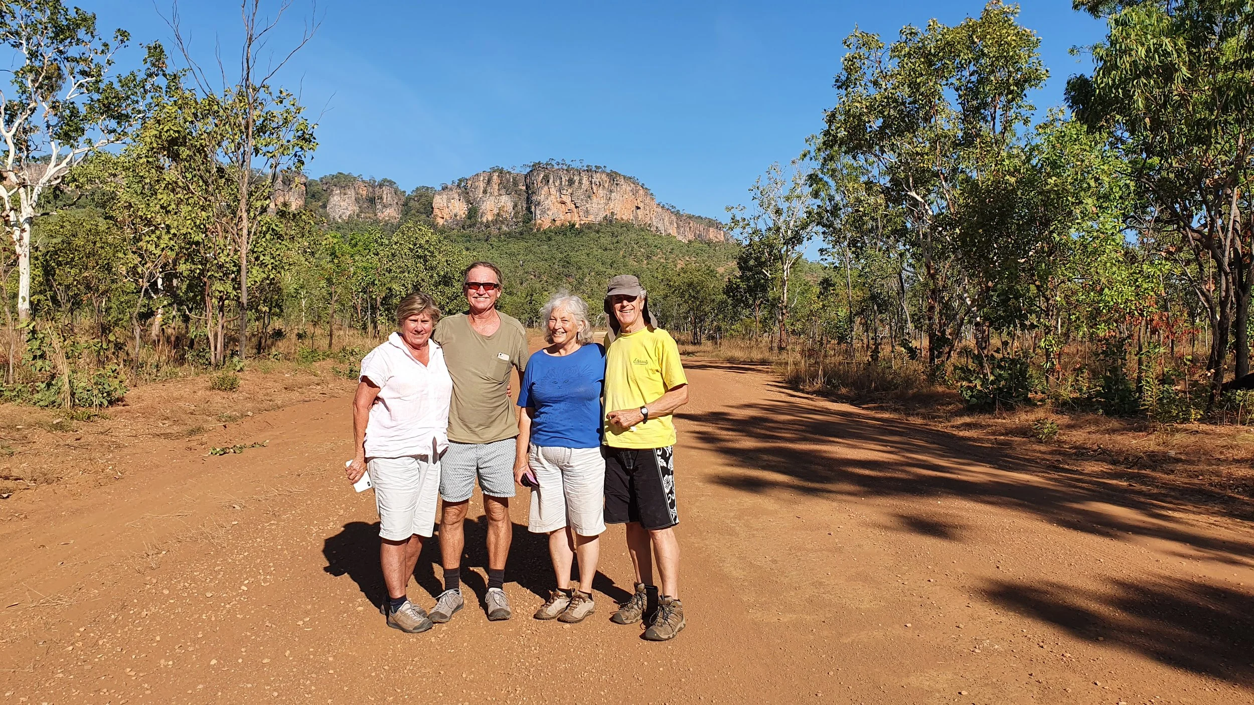 Four people standing on a dirt path in a forest with a large rocky mountain in the background, under a clear blue sky.