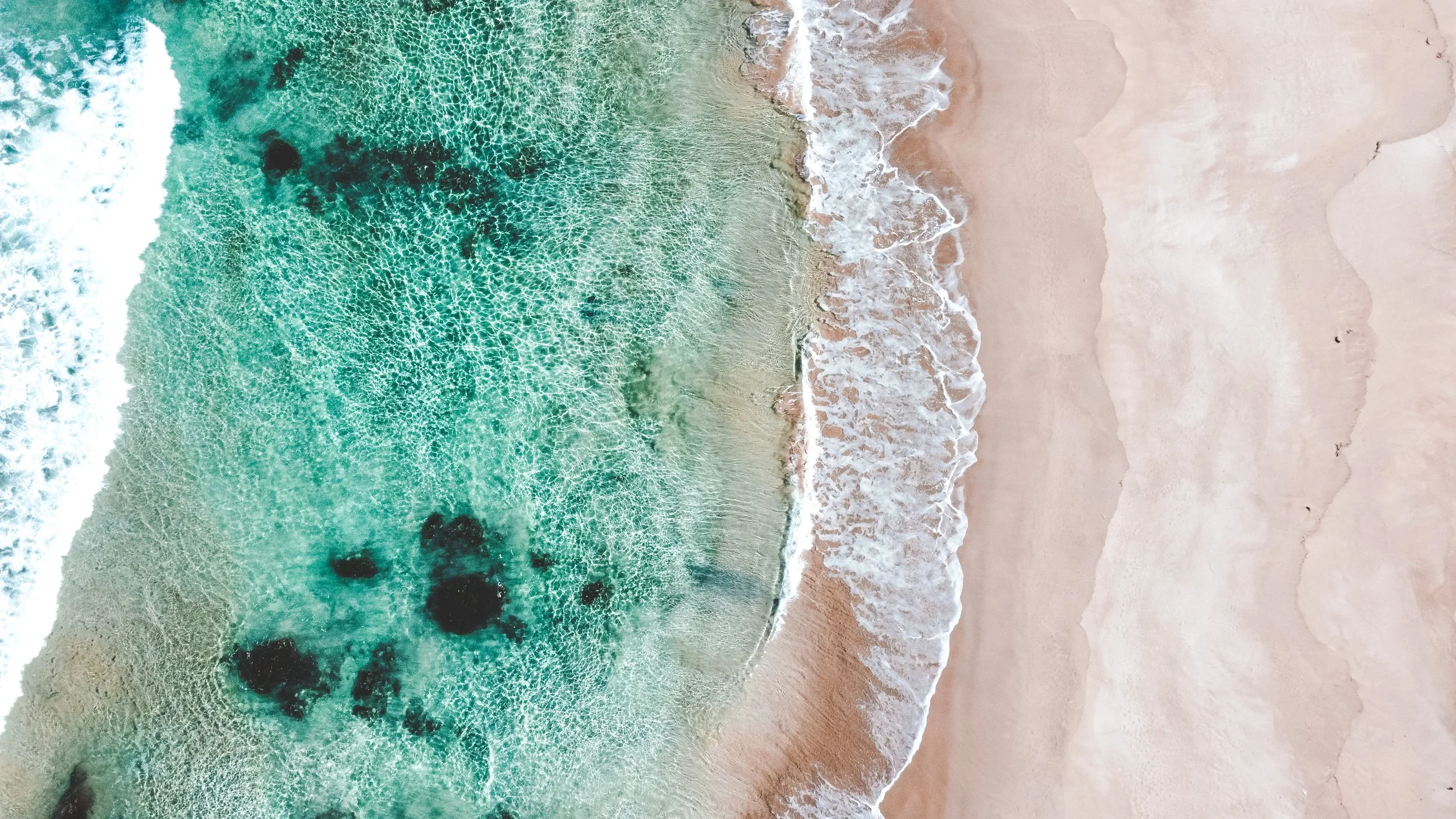 Aerial view of a beach with clear turquoise water and white sand, with waves gently washing onto the shore.