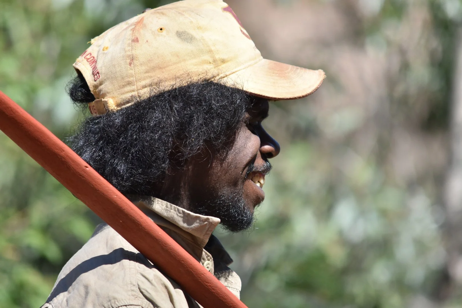 A man with dark skin and curly hair wearing a beige cap and beige shirt, smiling as he holds a red stick or pole, outdoors with blurred greenery in the background.