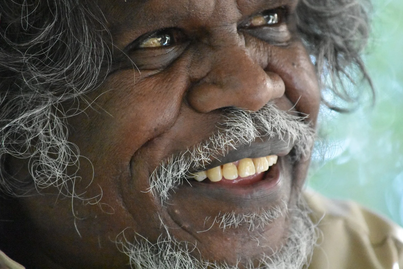 Close-up of a smiling elderly man with gray hair and beard, showing his teeth.
