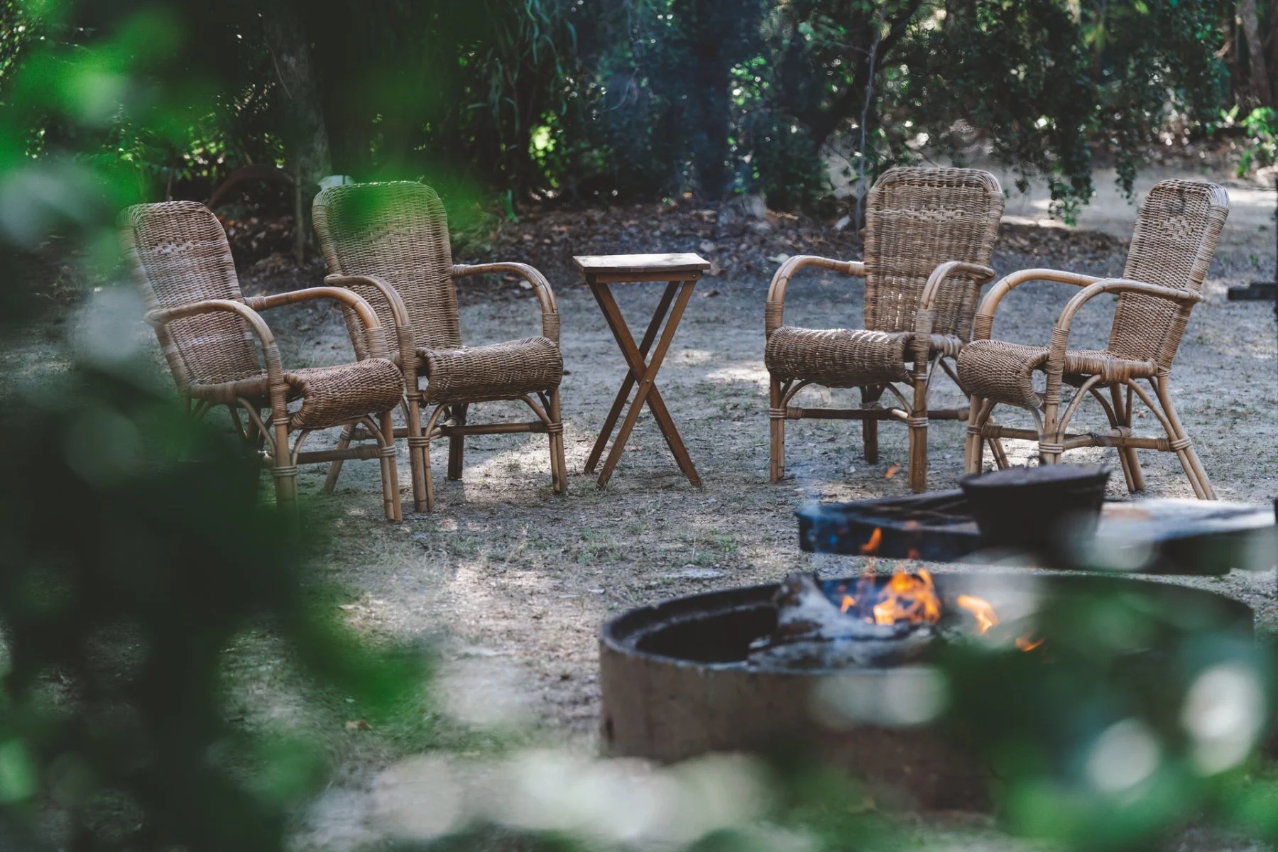 Group of four wicker chairs arranged around a small wooden table outdoors in a wooded area, with a fire pit in the foreground.