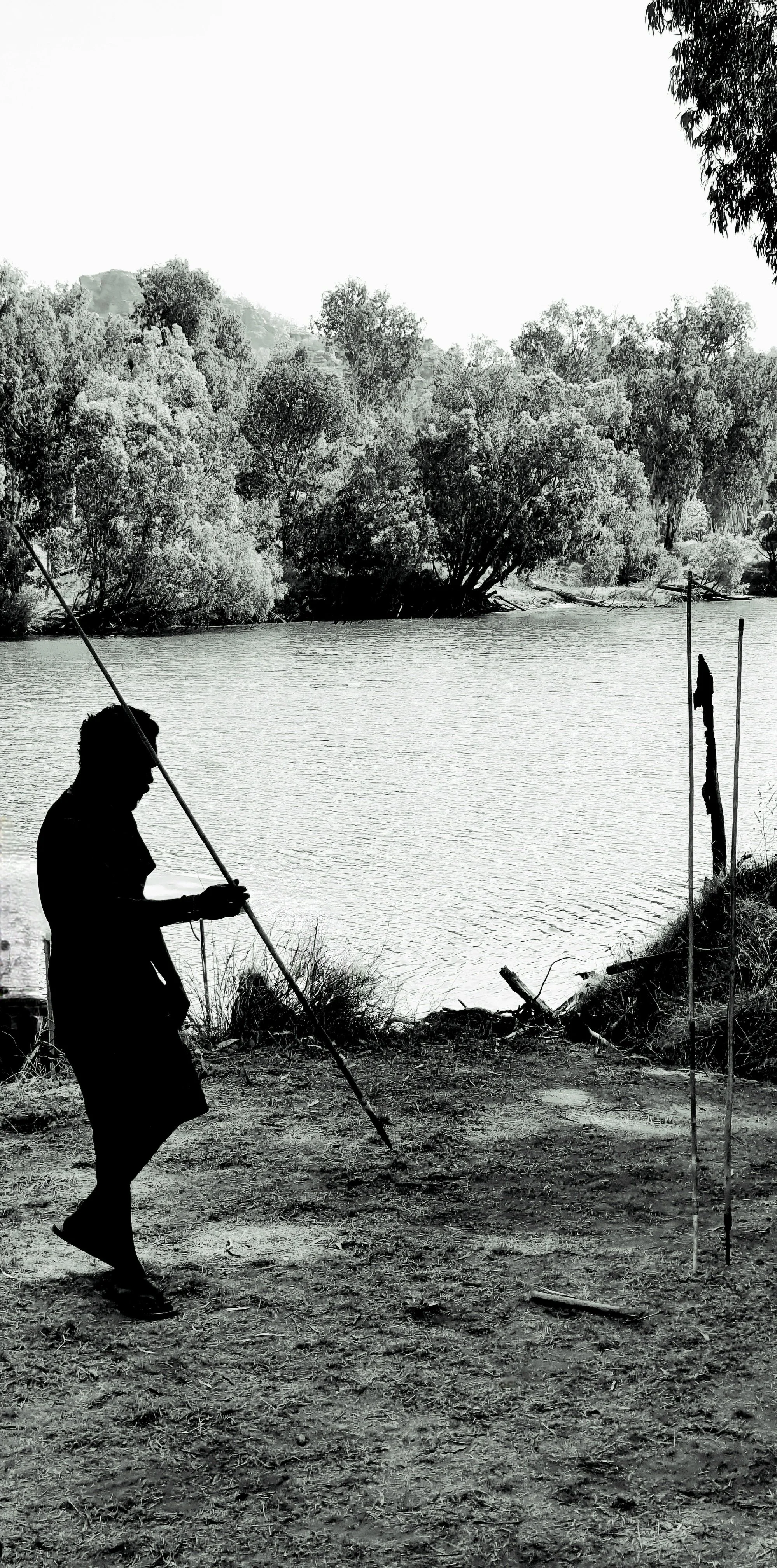 Silhouette of a person standing on the riverbank holding a stick, near a body of water with trees in the background.
