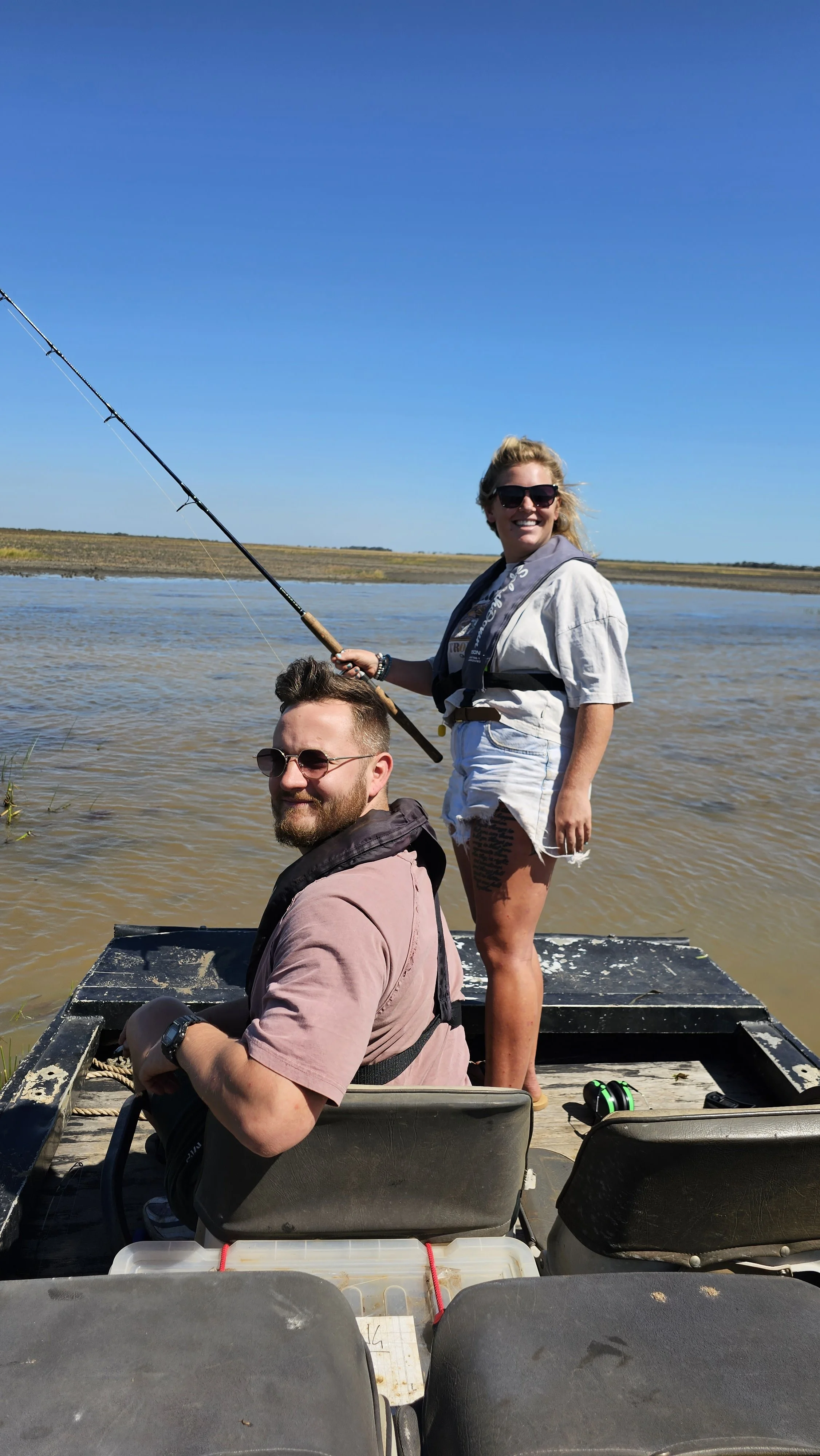 Two people fishing on a boat in a river or lake under a clear blue sky, with dry land visible in the background. One person is standing with a fishing rod, and the other is sitting, both smiling.