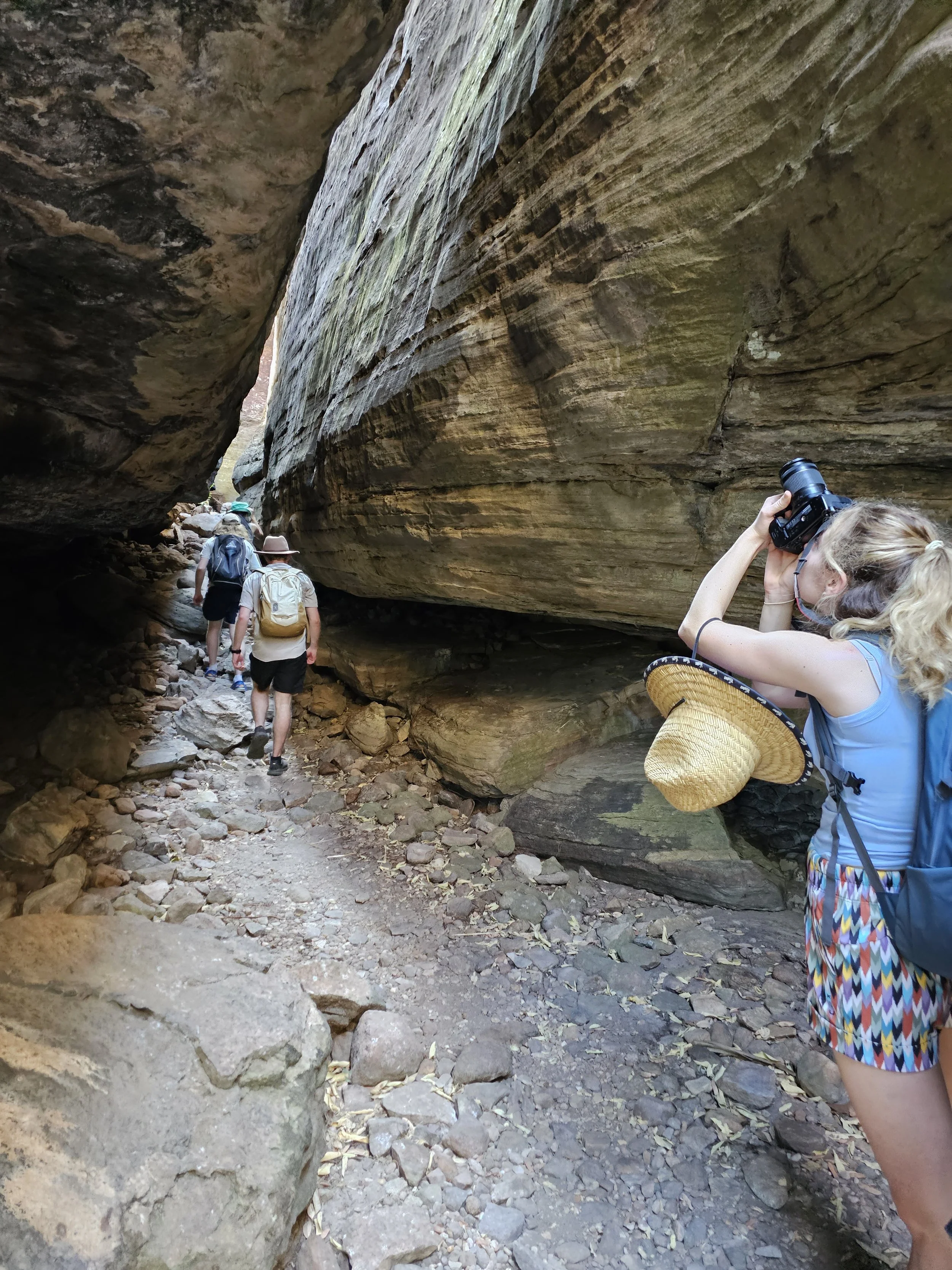 A group of hikers walking through a narrow canyon with high rock walls, some of which are layered and weathered. One woman in the foreground is taking a photo with a camera, wearing a hat and colorful shorts, while others walk ahead.