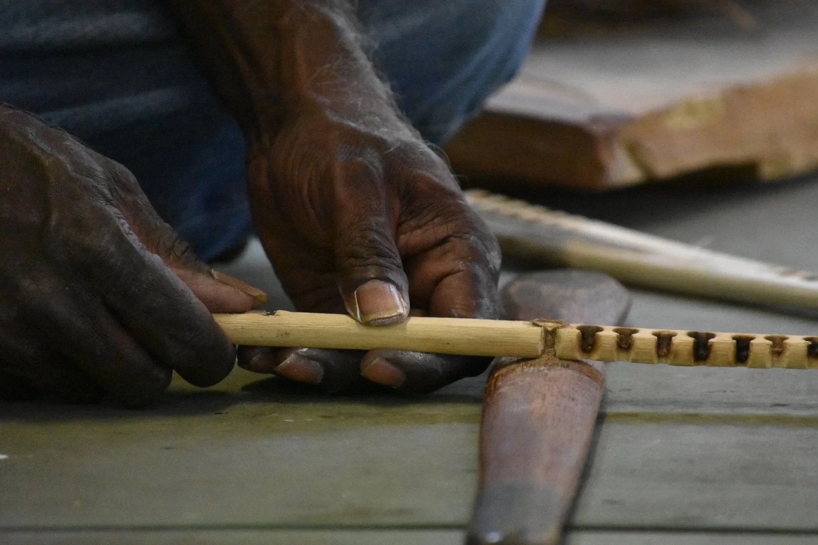 Close-up of hands working on a wooden object, possibly using a chisel or carving tool, with a workspace background.