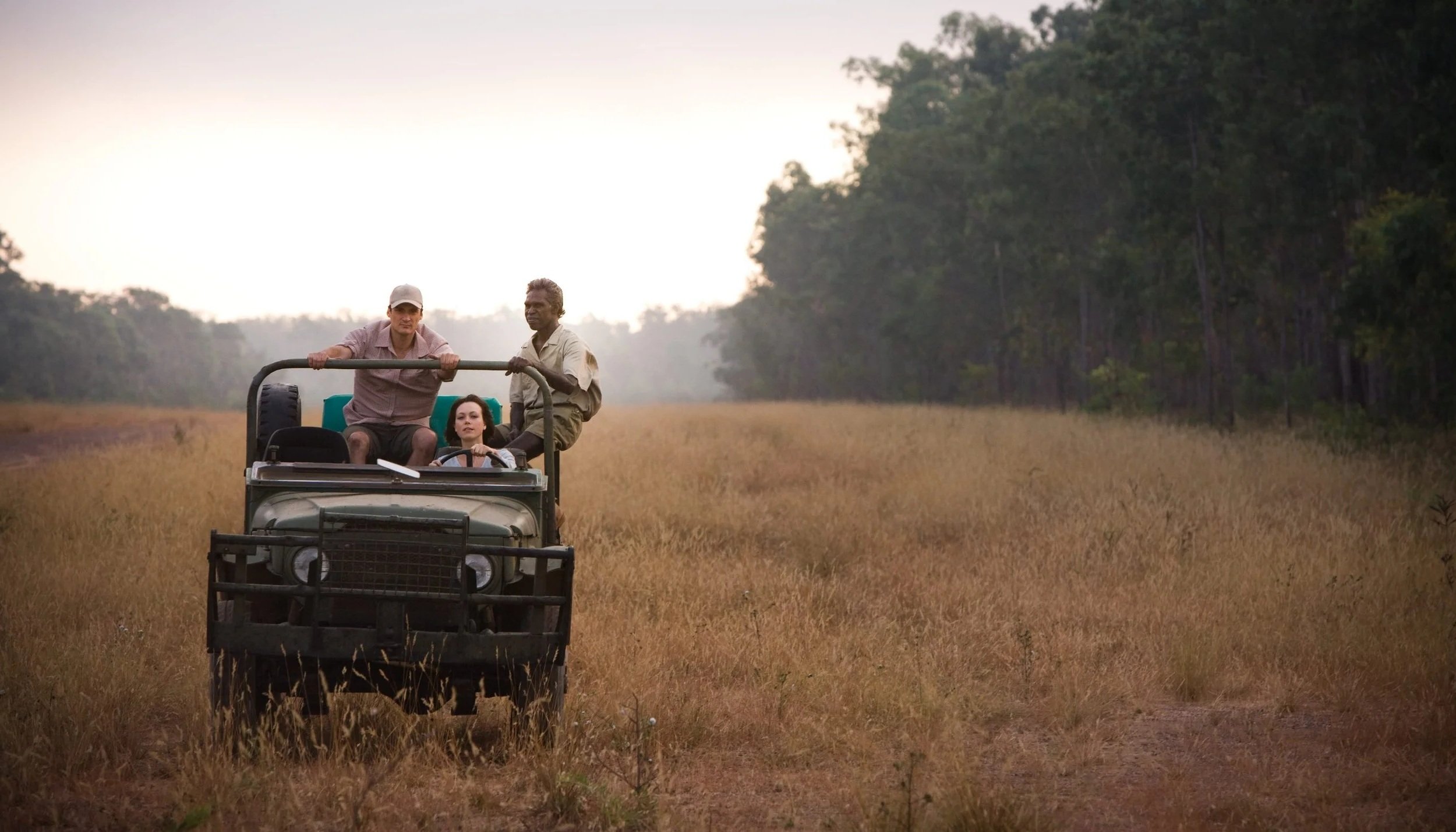 Three people riding an open vehicle on a dirt path through a grassy field with trees in the background.