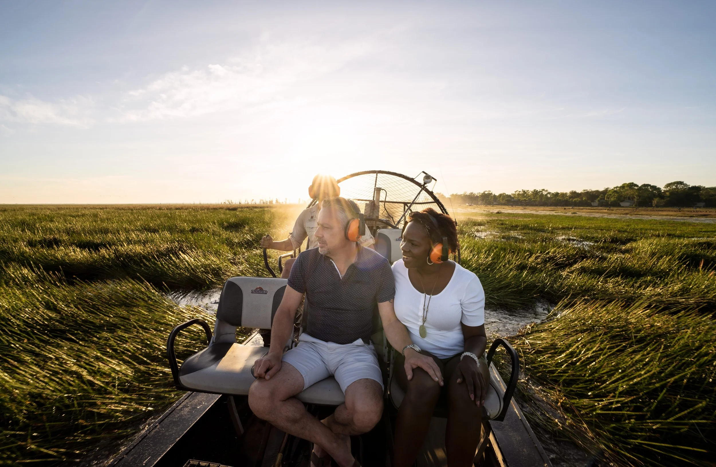 Two people sitting together on a golf cart in a field with tall grass at sunset, with a person in the back operating the cart, and trees in the distance.