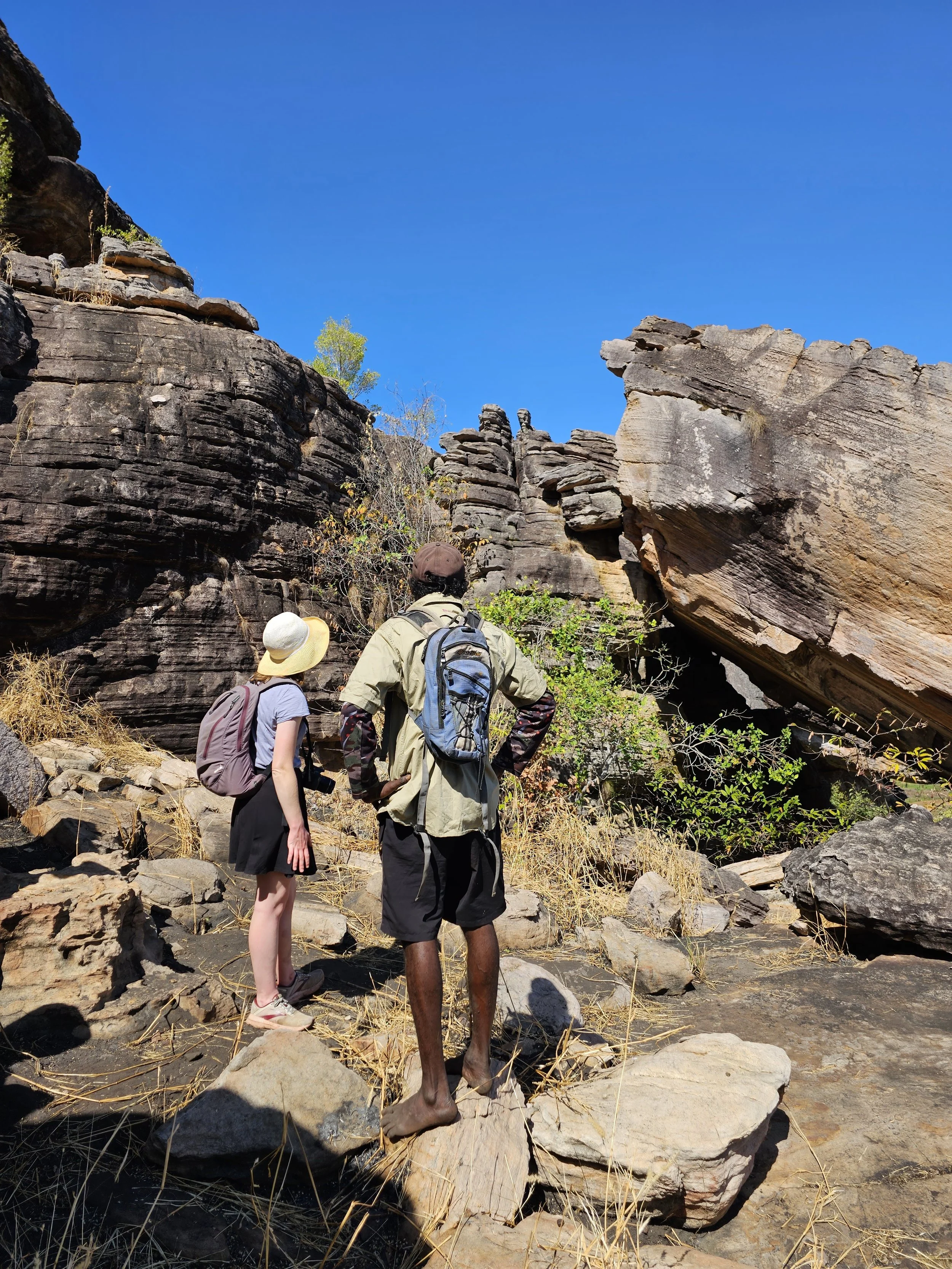 Three hikers standing on rocky terrain in a canyon, looking at large rock formations against a clear blue sky, with some vegetation and small trees around.