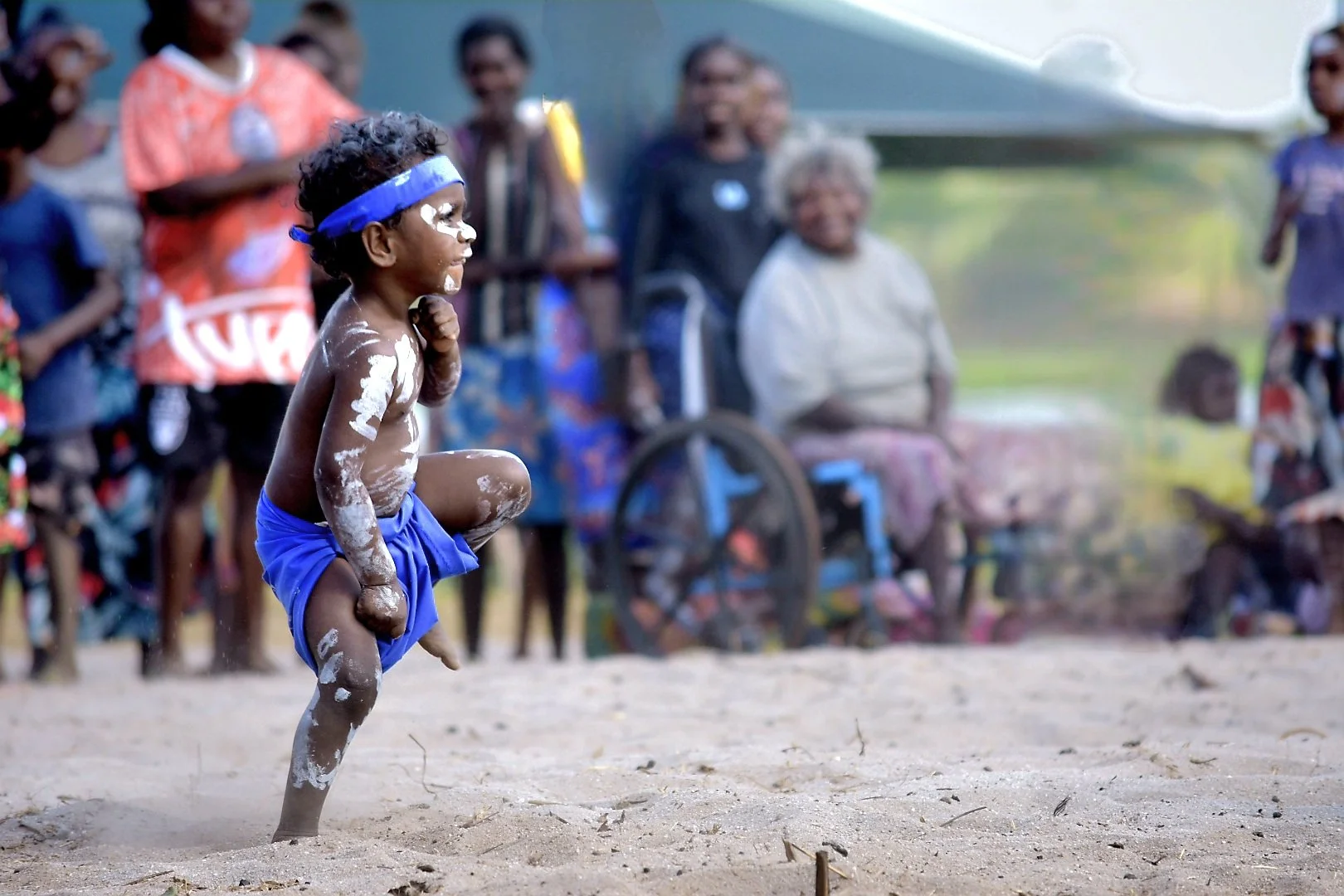 A young boy running on a sandy field during a traditional event, wearing blue shorts and a blue headband, with white paint on his face and body, while a crowd of onlookers observes in the background.
