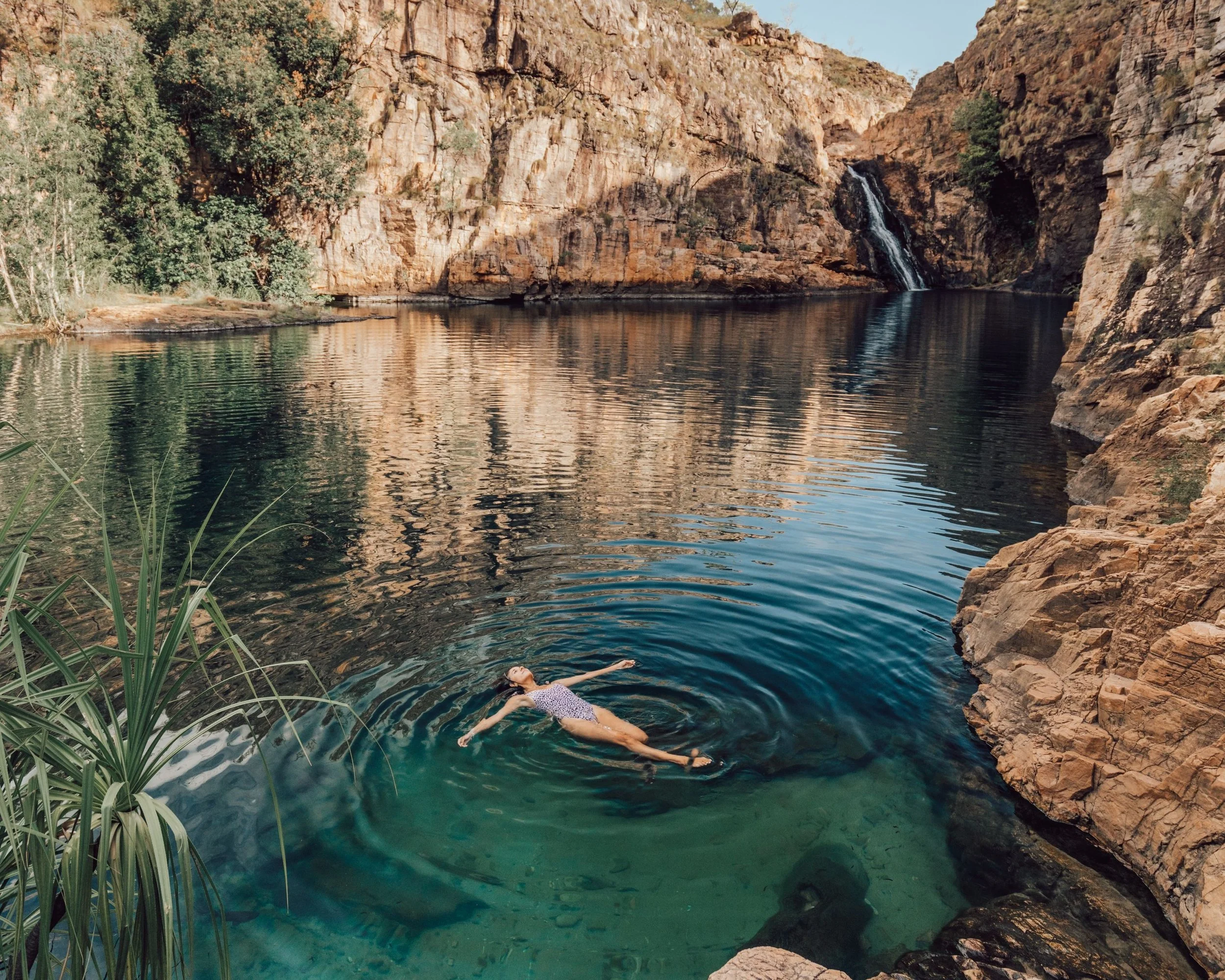 A woman in a swimsuit floating on her back in a calm river surrounded by rocky cliffs and greenery, with a small waterfall in the background.