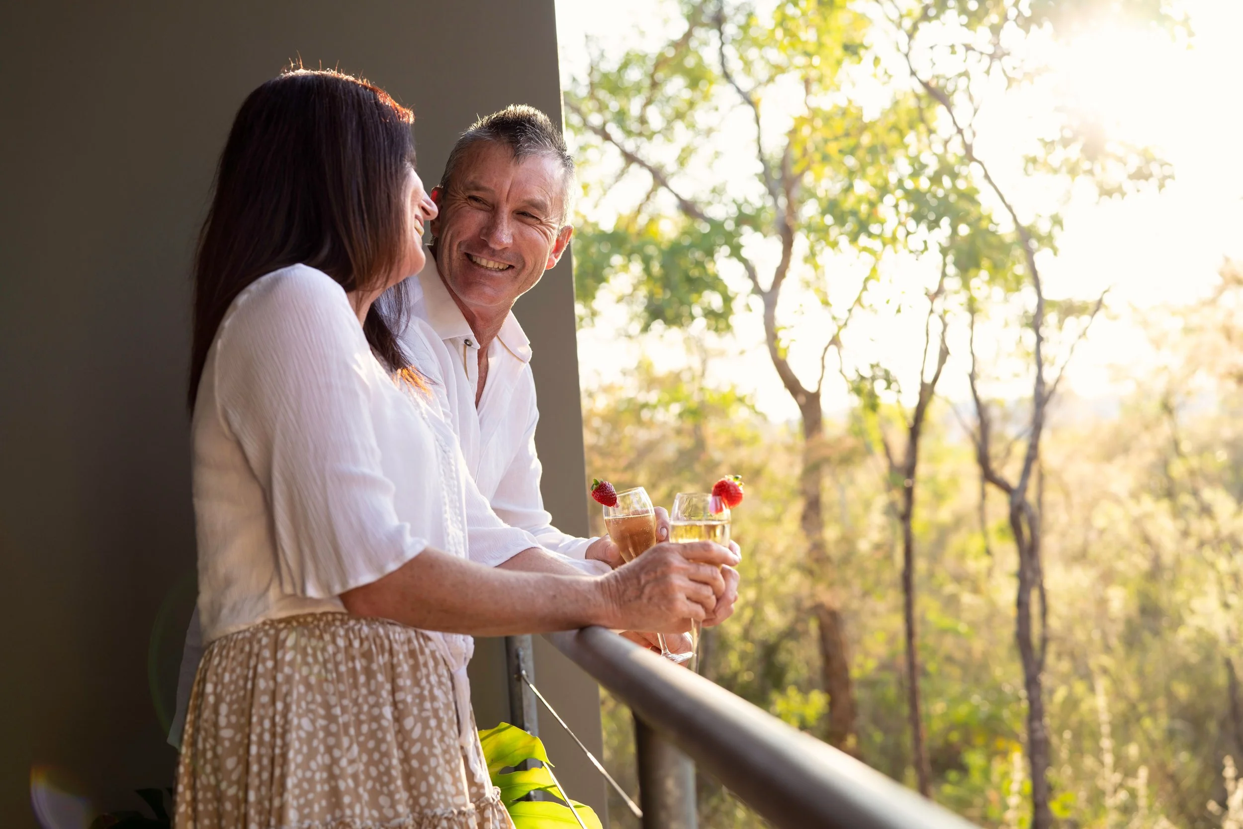 Two people enjoying drinks and conversation on a balcony with trees and sunlight in the background.