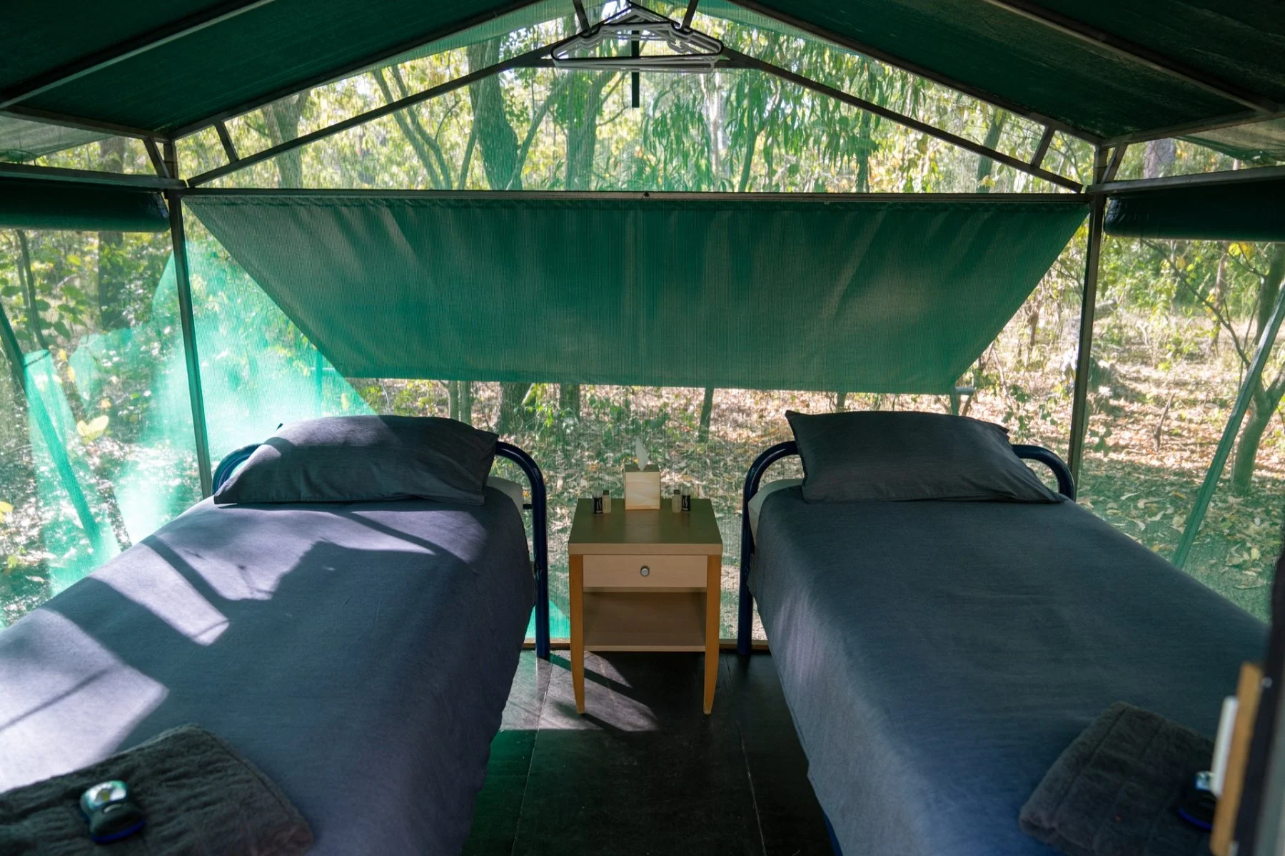 Two twin beds with dark gray sheets and pillows in a screened outdoor room surrounded by leafy green trees, with a small wooden nightstand between them holding a tissue box and small bottles.