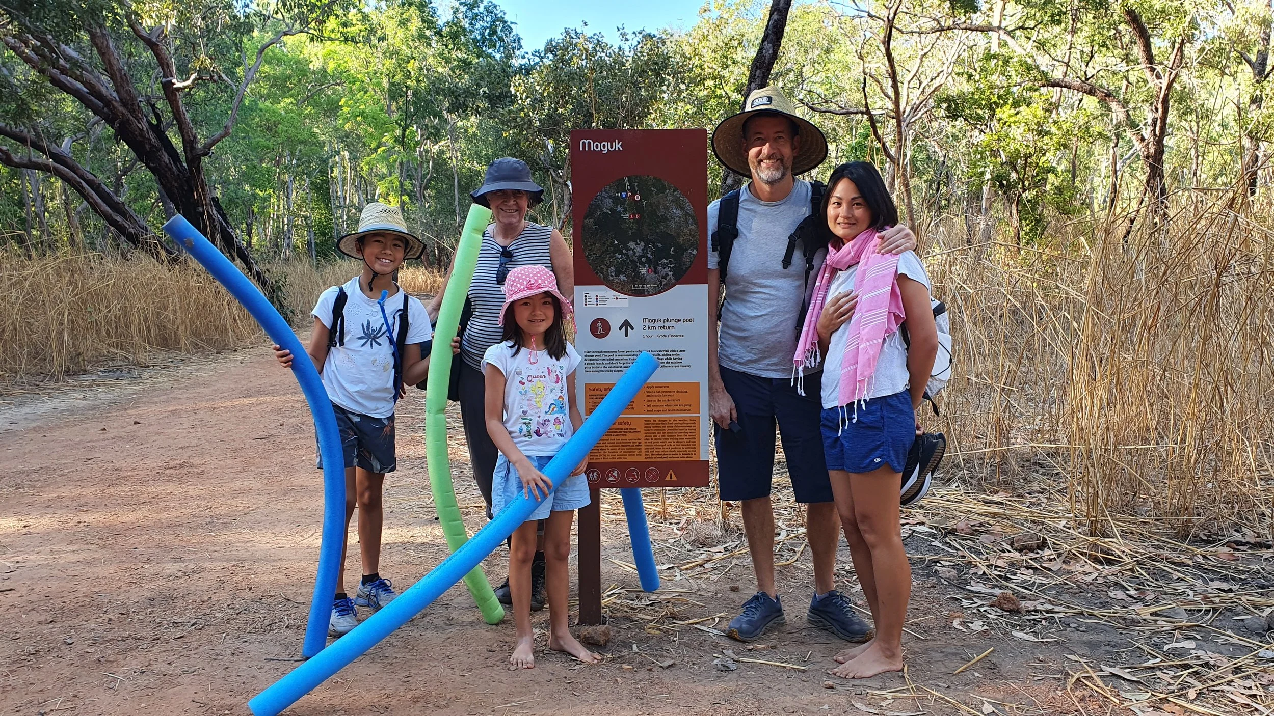 Group of five people standing outdoors near a sign about Maguk. They are dressed for a day in the sun, with some wearing hats and backpacks. Two children hold pool noodles, and all are smiling. The background features trees and dry grass on a dirt pa