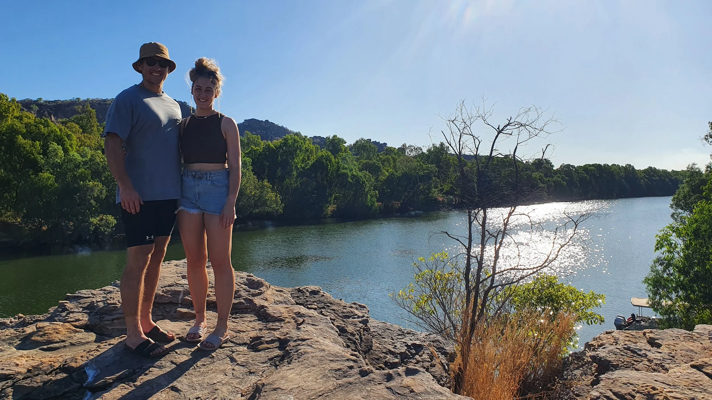A man and a woman standing on a rocky shore by a river, smiling at the camera with trees and a mountain in the background on a sunny day.