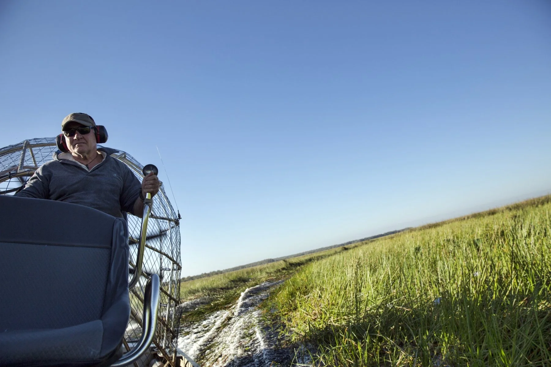 A man wearing headphones and sunglasses controls a hot air balloon's burner over a grassy field with a small water stream, under a clear blue sky.