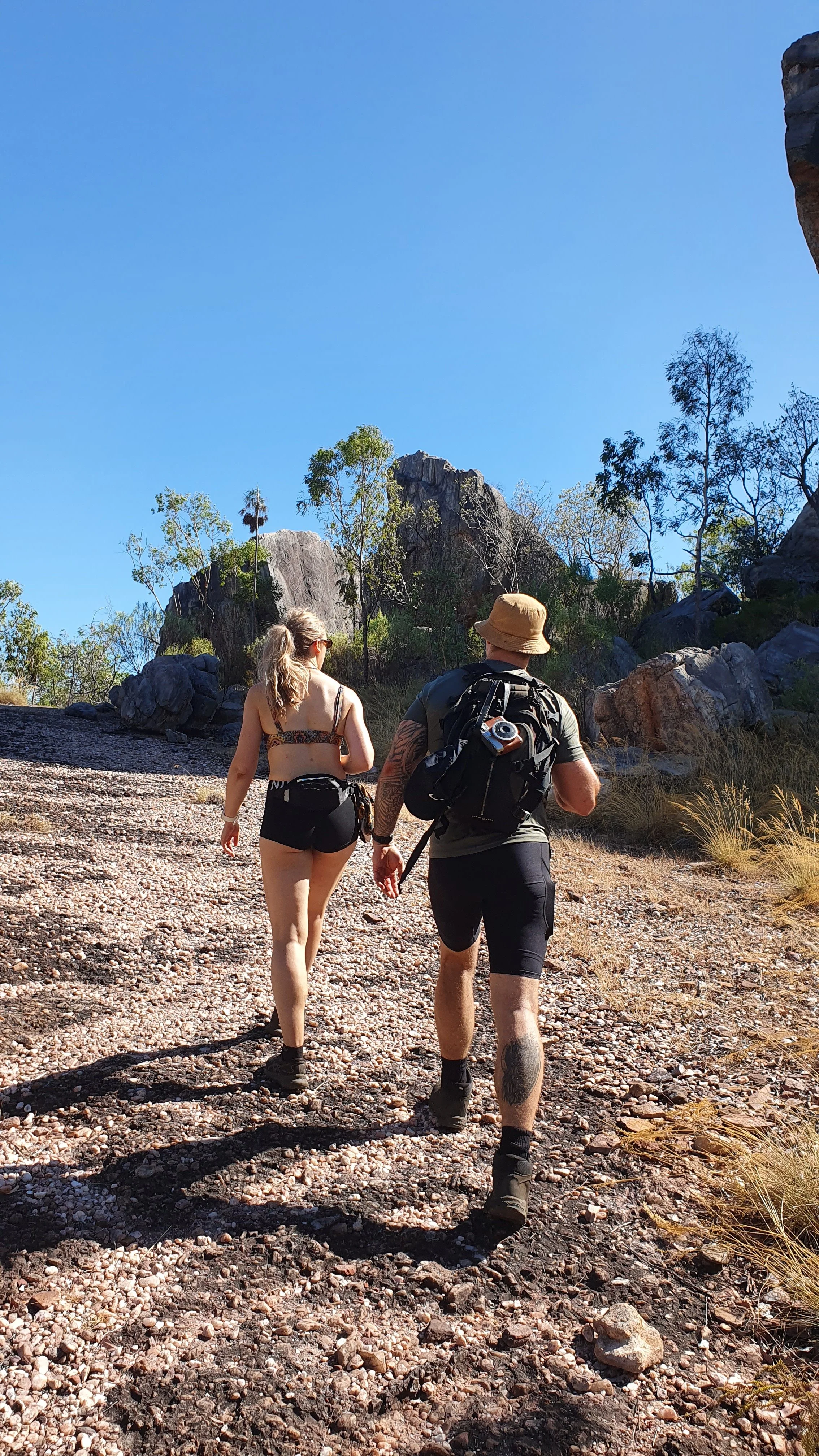 Two people hiking in a rocky, desert landscape with large rocks and sparse trees under a clear blue sky.
