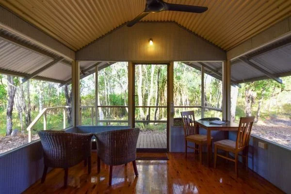 Sunroom with wooden floors, wicker chairs, a small table, and sliding glass doors overlooking a wooded area.