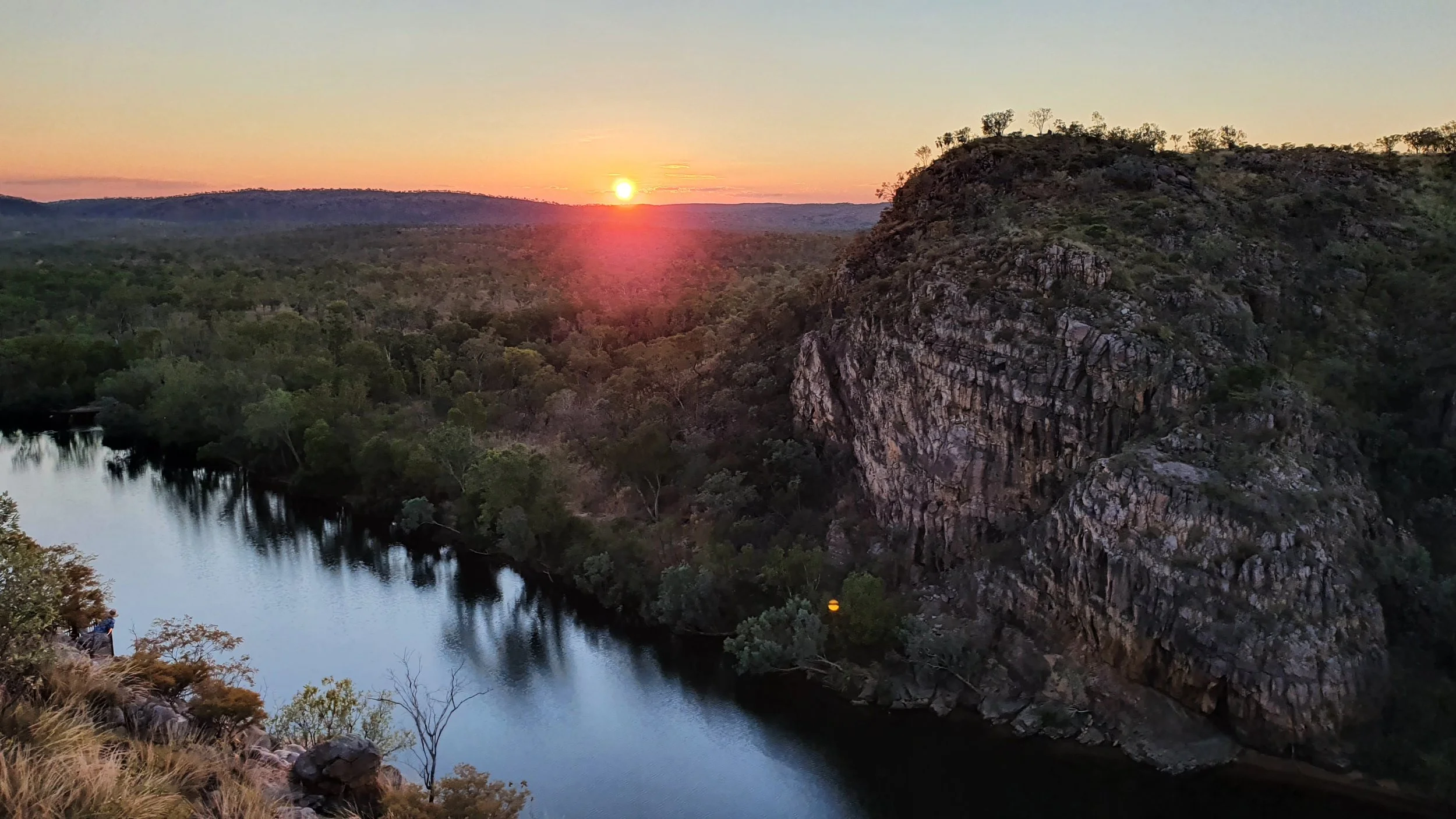 Sun setting over a river with surrounding trees and rocky hills in the distance.
