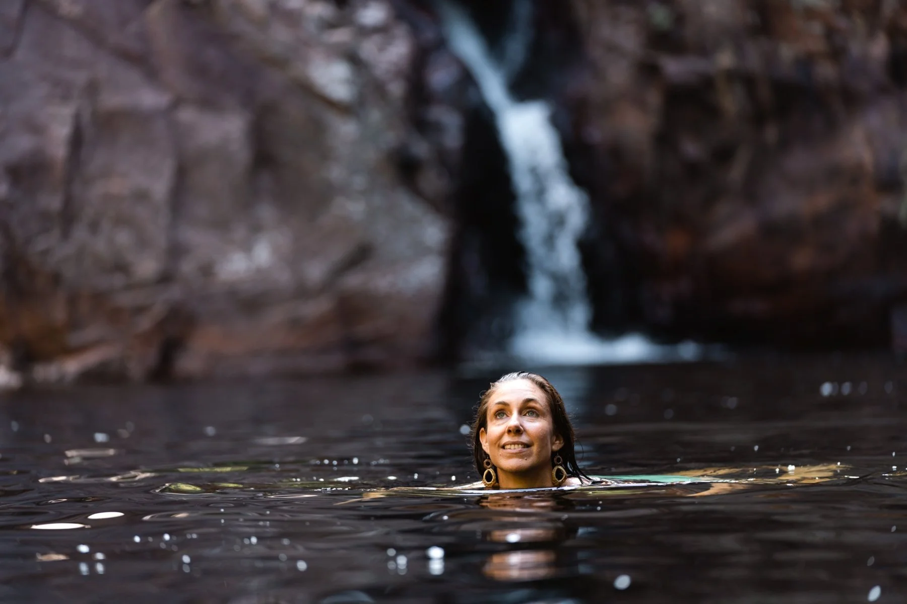 A woman with dark hair and earrings swimming in a natural water pool with a waterfall in the background.