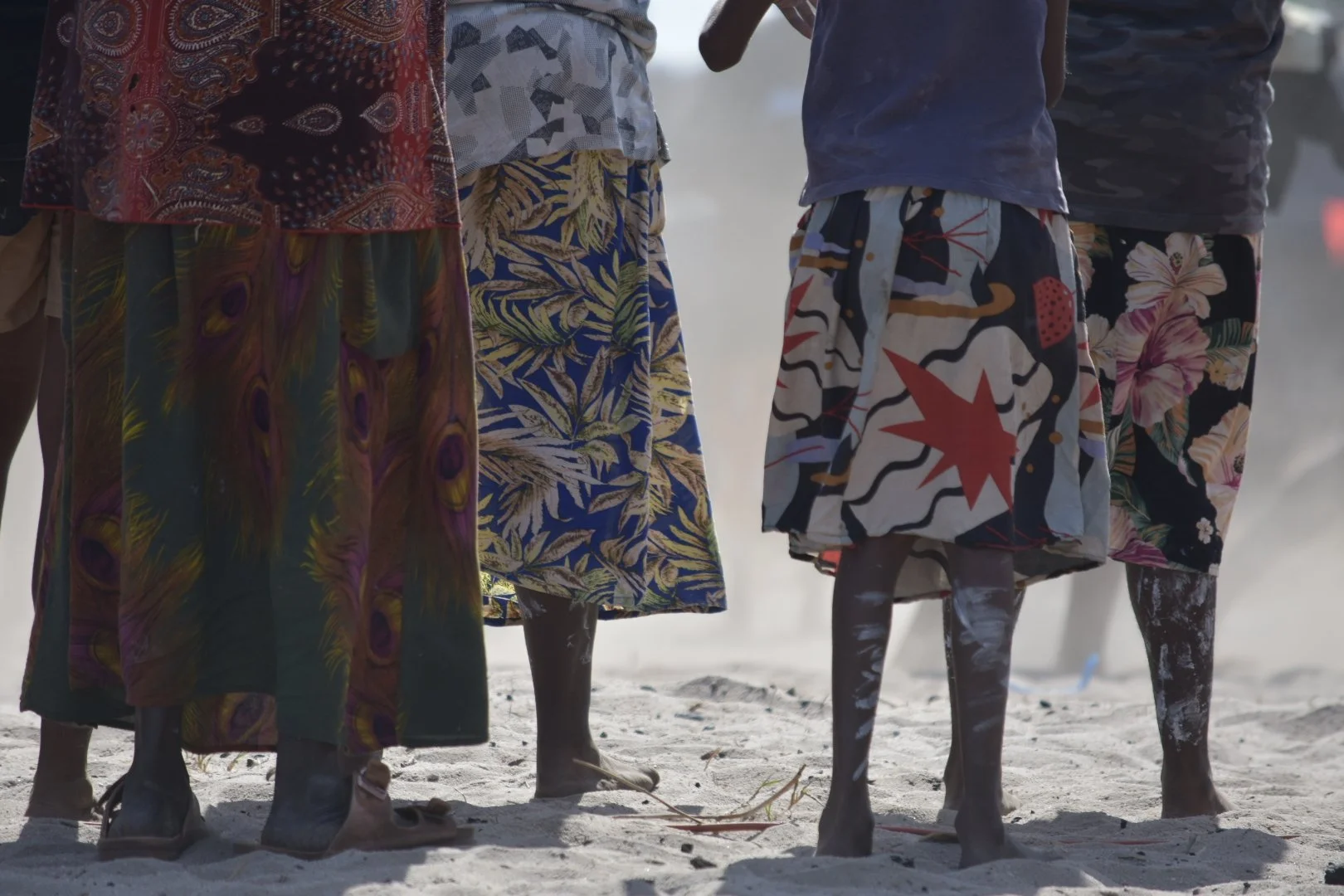 Group of people standing on sandy ground, wearing colorful, patterned skirts and shorts, with their legs visible from the knees down.