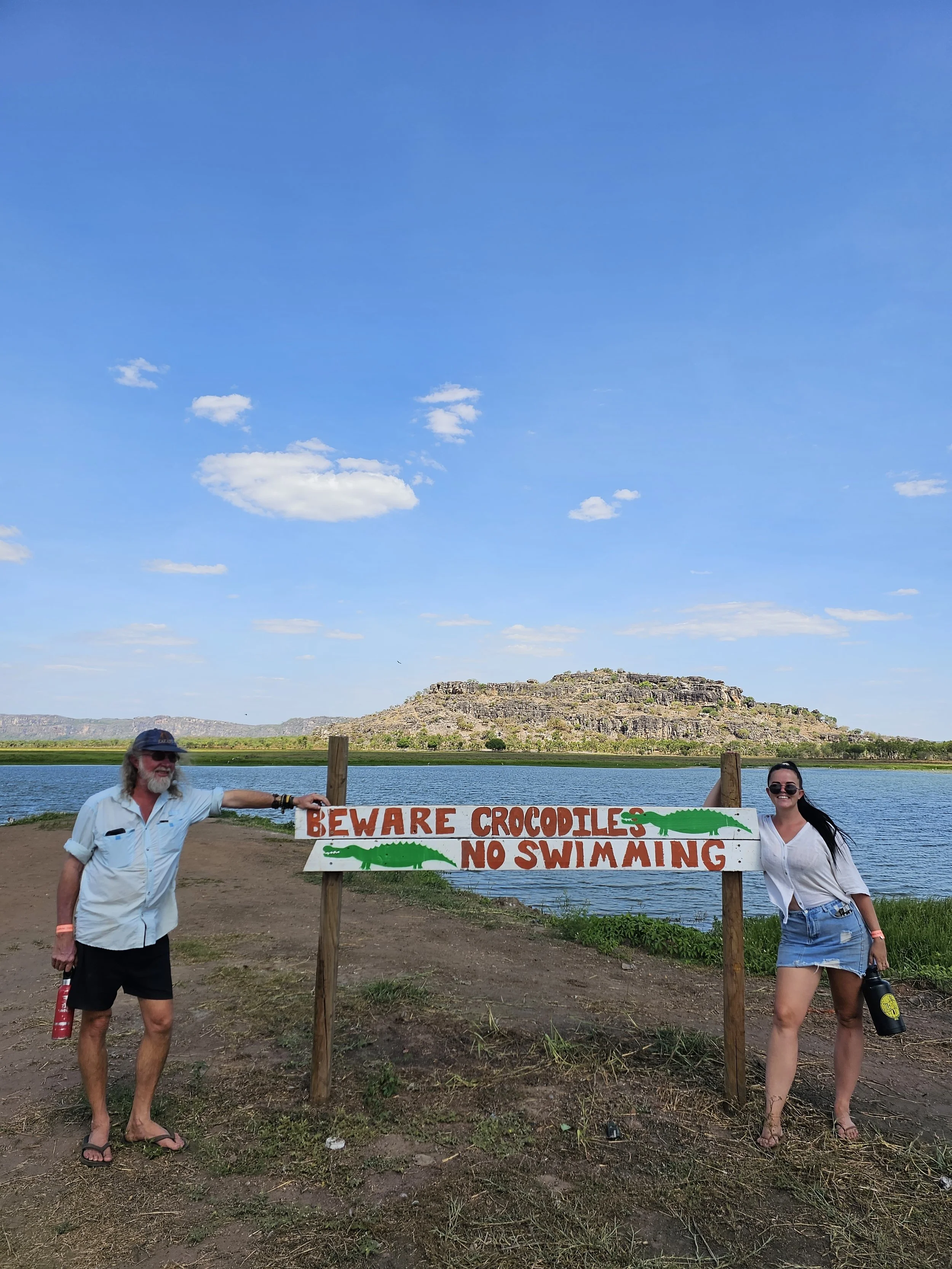 Two people standing on either side of a sign that reads "Beware Crocodiles No Swimming" near a body of water with a large rocky hill in the background. The person on the left is an older man with gray hair, wearing a white shirt, black shorts, flip-f