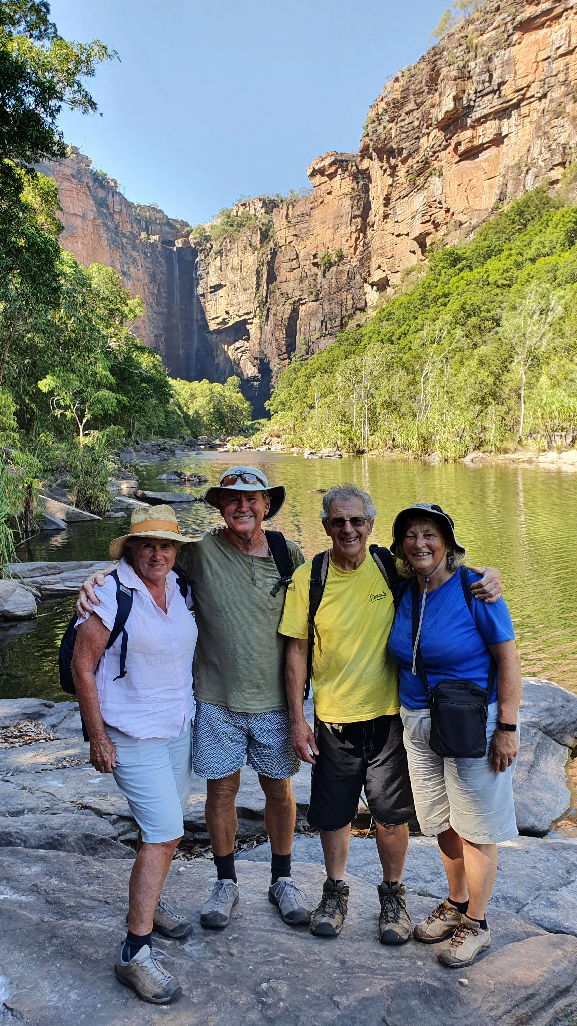 Four seniors standing on rocks beside a river, with a waterfall and tall cliffs in the background, smiling and dressed in hiking clothes and hats.