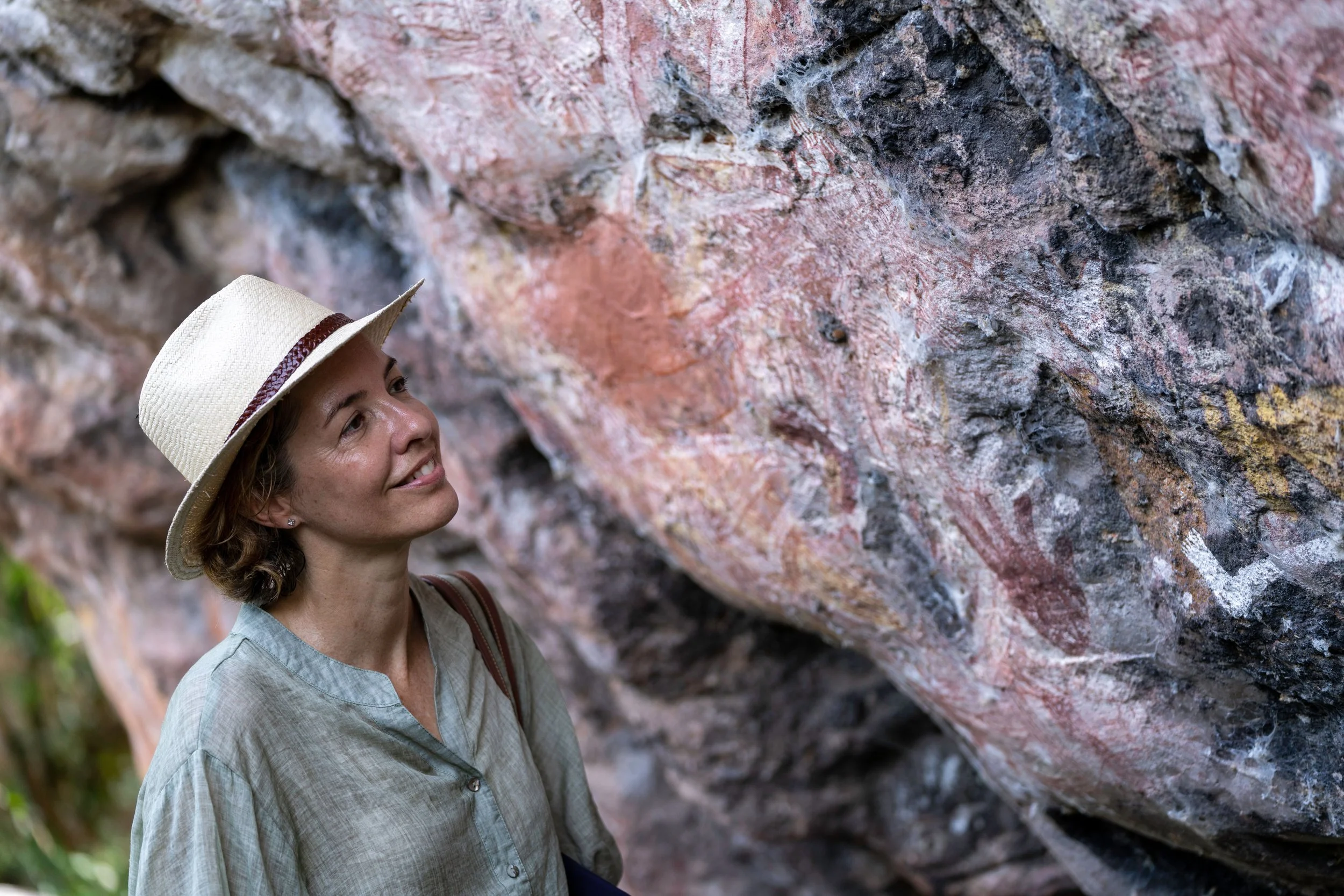A woman with a hat smiling while standing near a large rock wall outdoors.