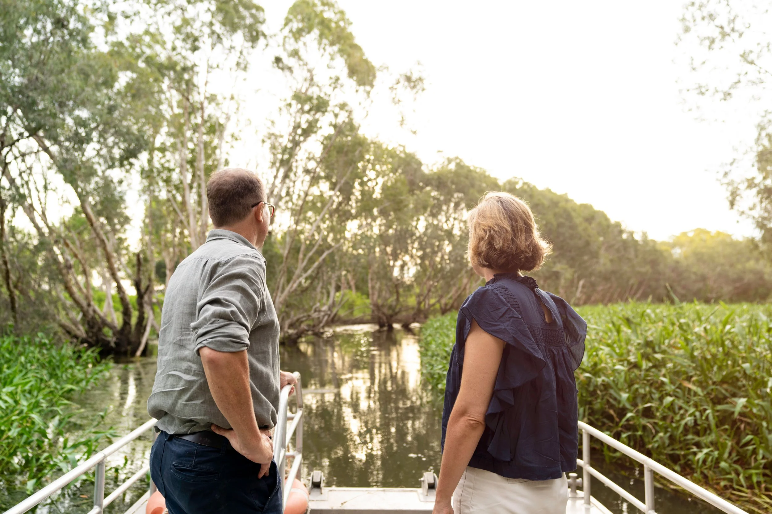 Two people standing on a bridge over a river, surrounded by lush green trees, with the sun setting or rising in the background.